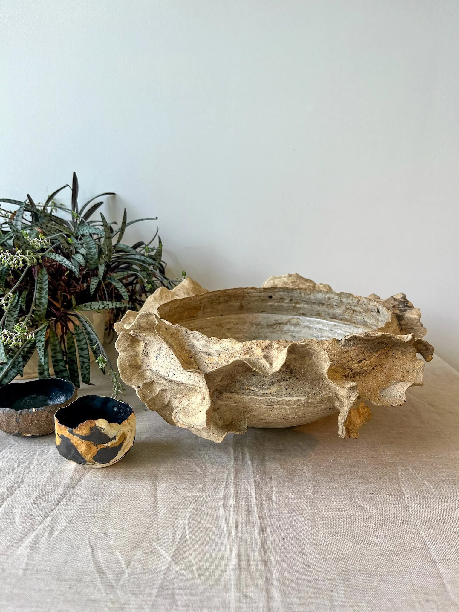 A large decorative bowl with a ruffled, natural stone-like appearance, flanked by two smaller black and gold ceramic bowls, placed on a beige tablecloth with a plant in the background against a plain wall.