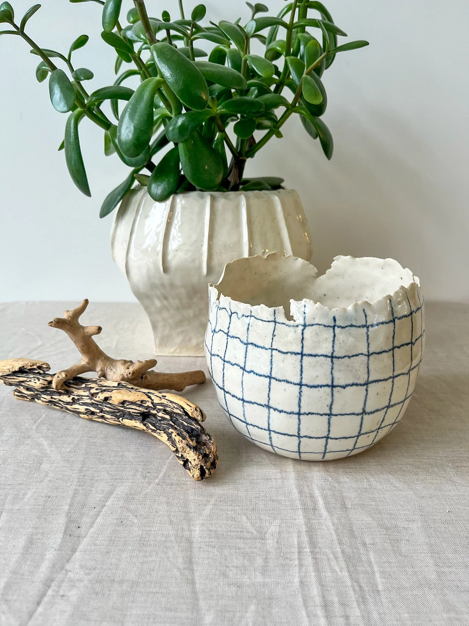 Decorative arrangement with a green potted plant, a partially broken ceramic bowl with a checkered pattern, a piece of driftwood, and a small wooden figurine on a neutral-colored tablecloth.