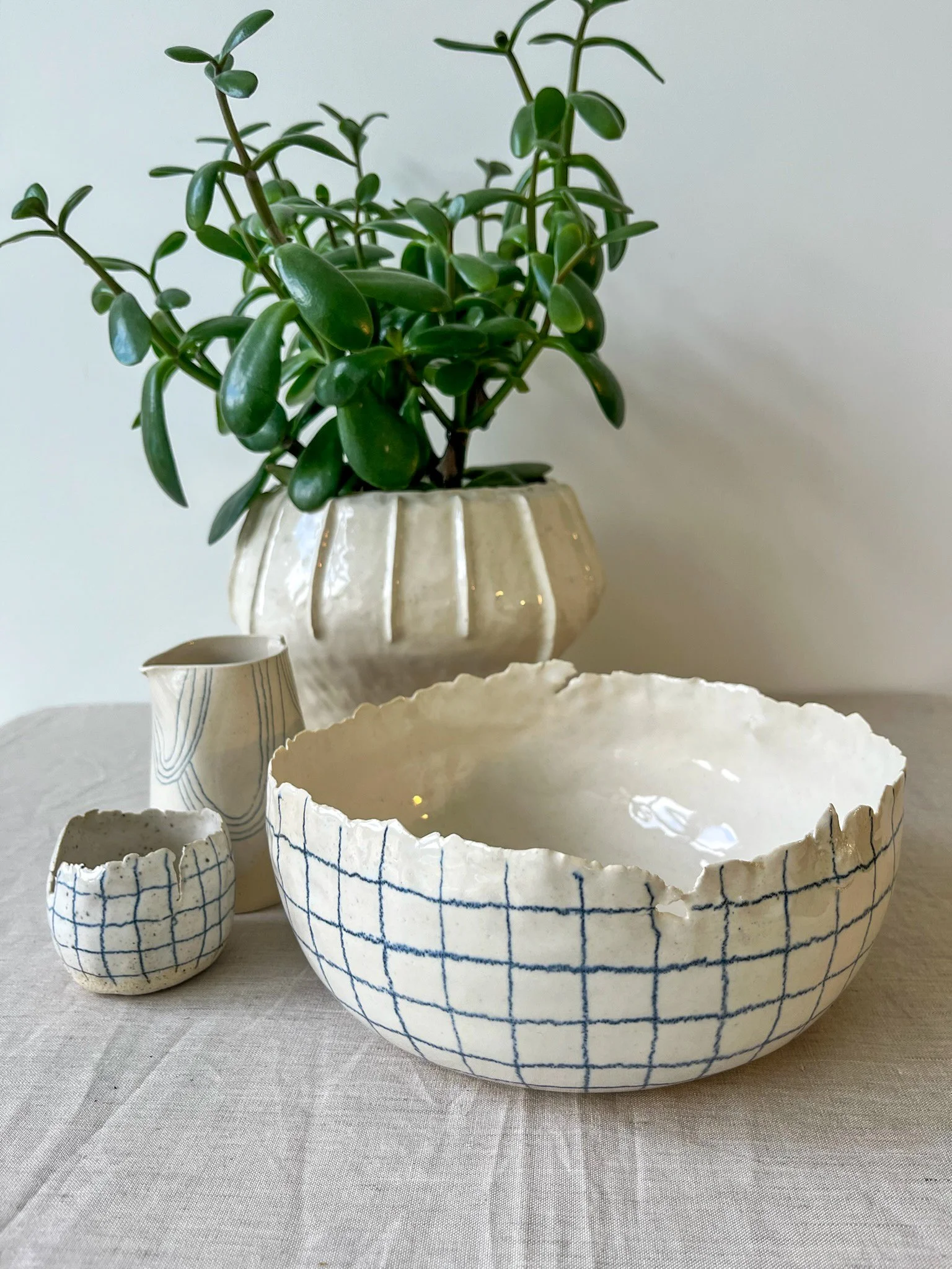 Decorative ceramic bowls and a small pitcher, with a potted jade plant in the background, on a neutral fabric surface.