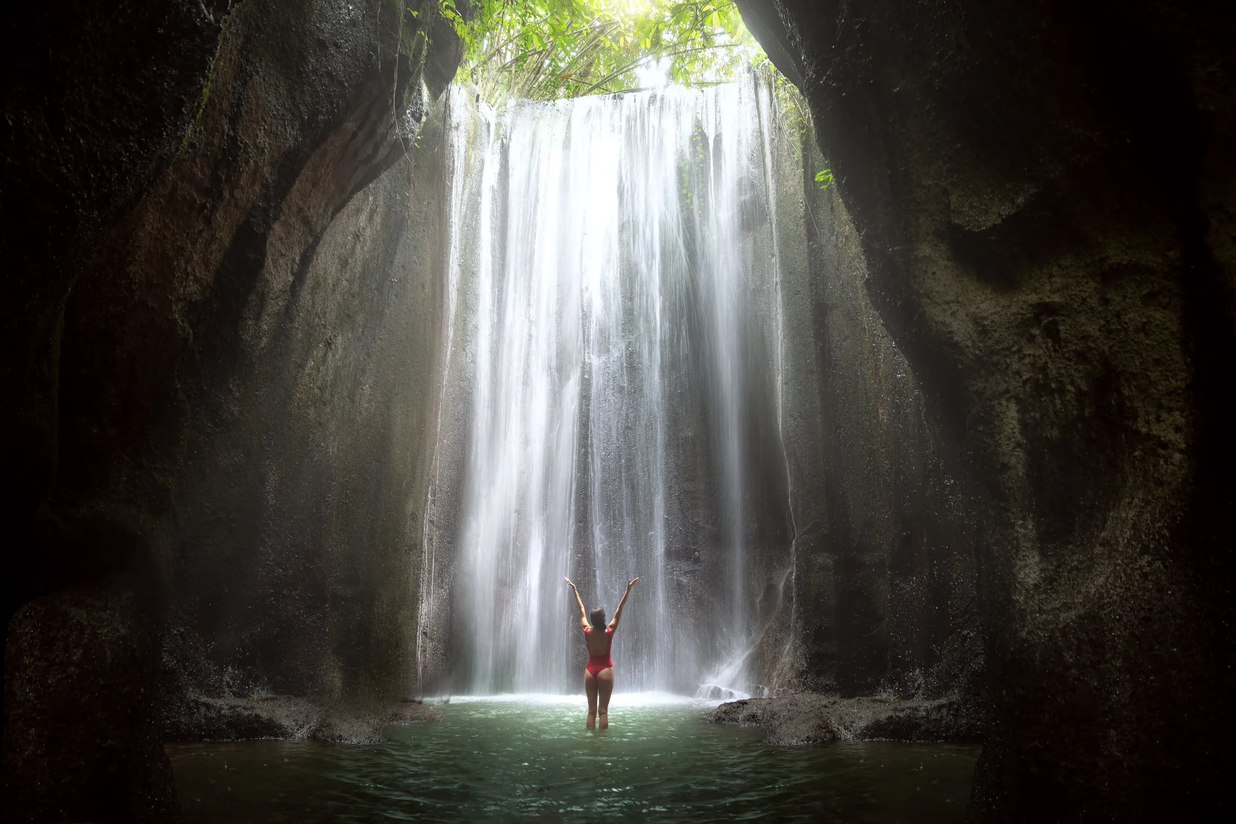 Woman with arms raised to gorgeous scenic epic majestic waterfall in cave with light rays