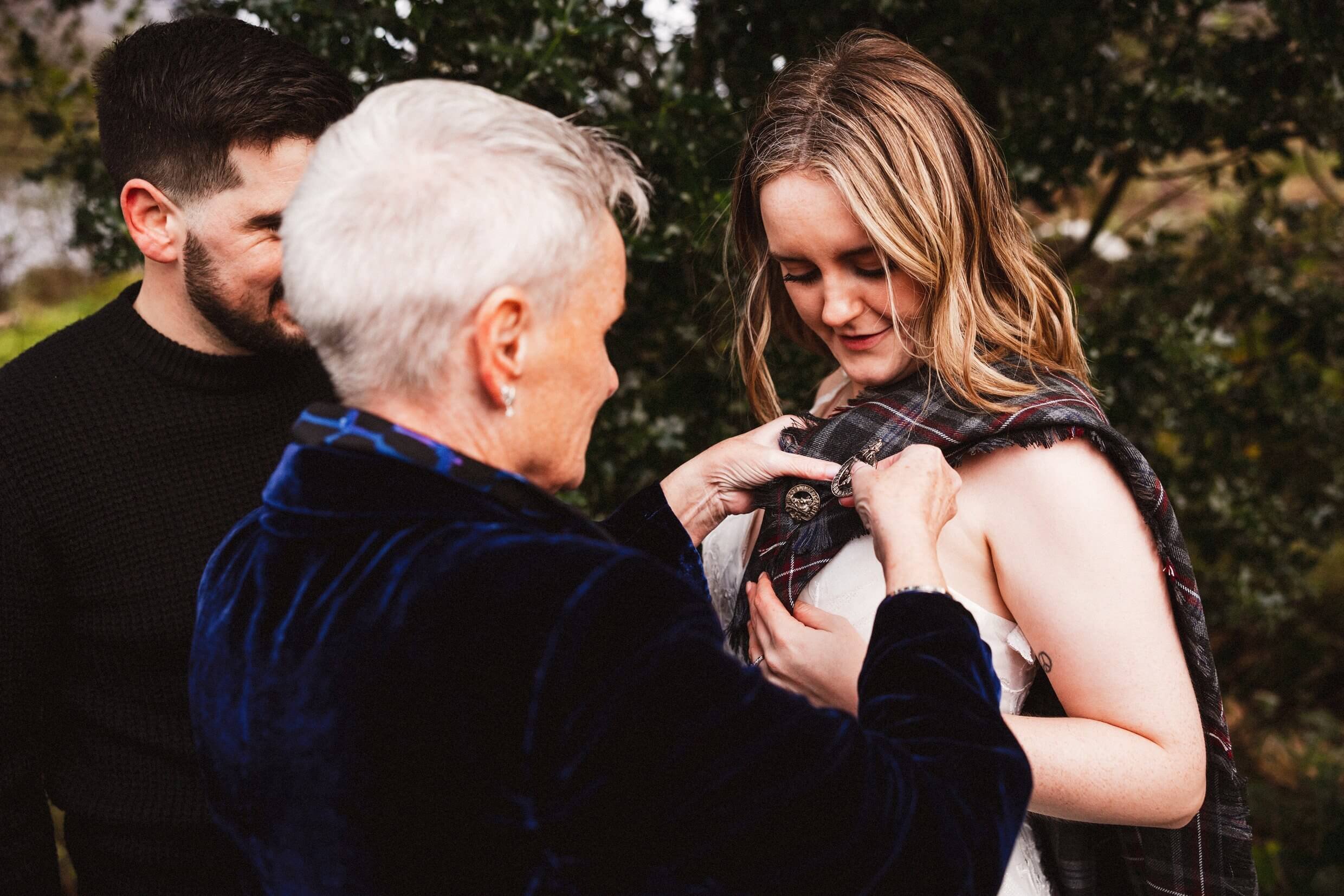 mother of the groom pins tartan plaid onto bride during scottish outdoor elopement in glencoe