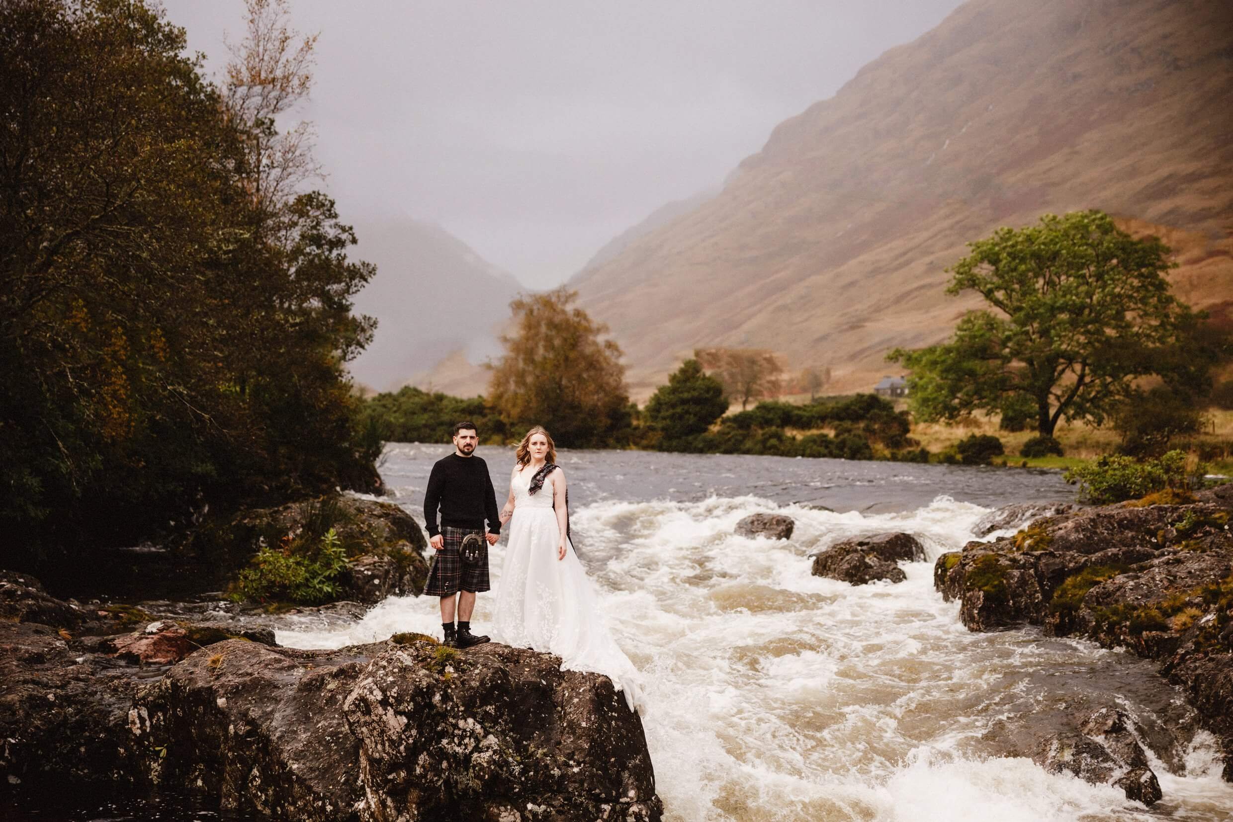 bride and groom stand next to river etive in glen etive following their glencoe elopement wedding ceremony in scotland