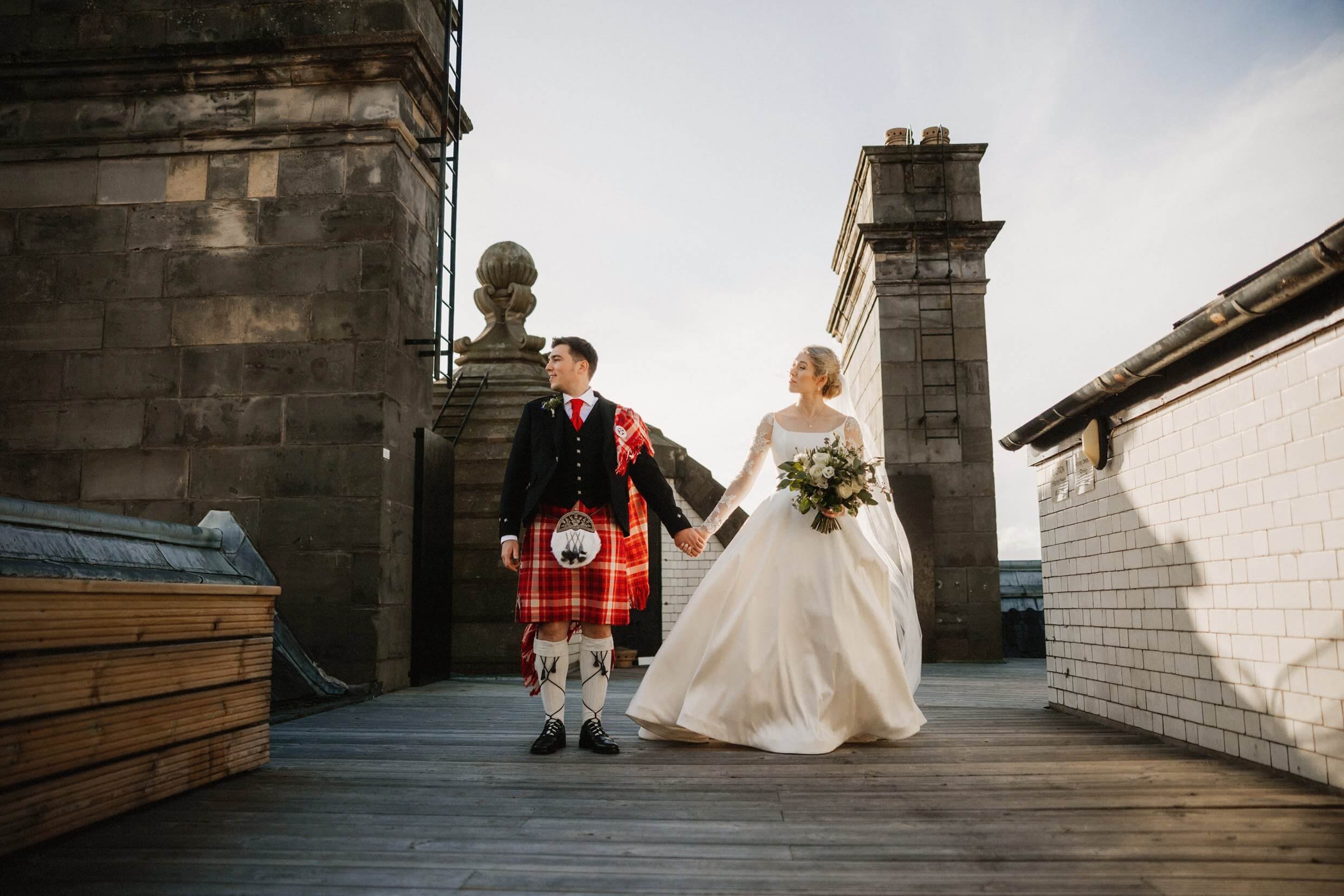 the bride and groom pose for photos in front of chimneys on the rooftop of the balmoral hotel edinburgh wedding venue in scotland