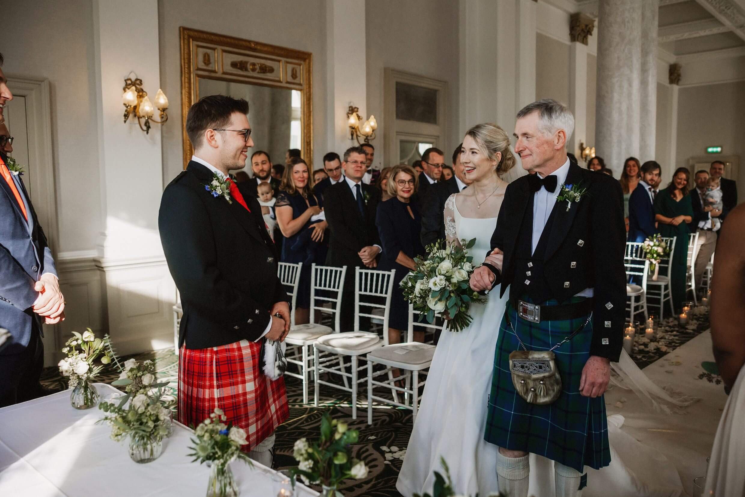 the bride and father of the bride meet the groom at the top of the aisle as guests look on during balmoral hotel edinburgh wedding ceremony in scotland