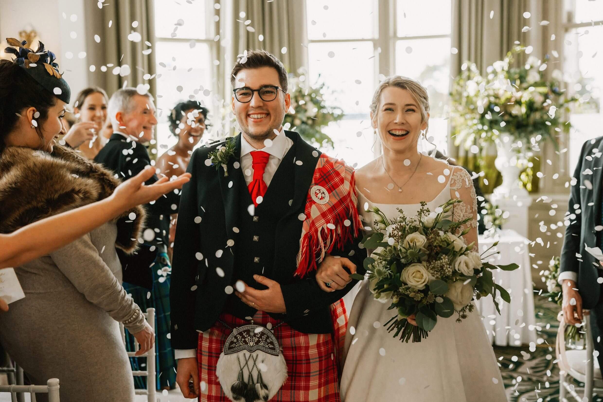 the bride and groom smile and link arms as guests throw confetti following a balmoral hotel edinburgh wedding ceremony
