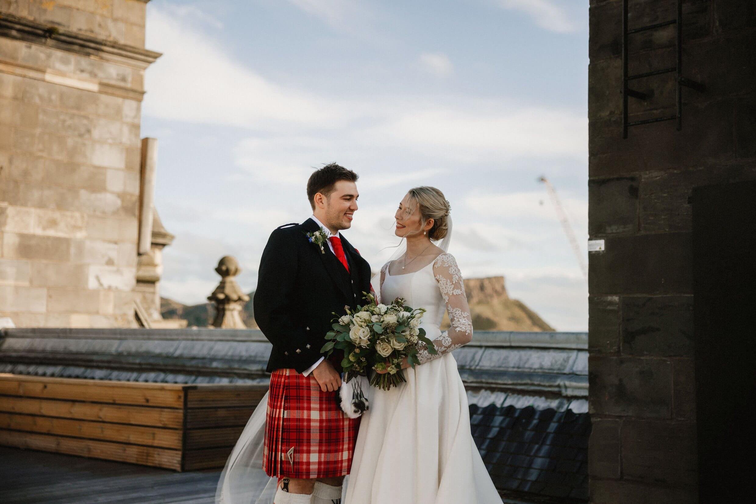 the bride and groom pose for photos on the rooftop of the balmoral hotel edinburgh wedding venue with arthur's seat visible in the background