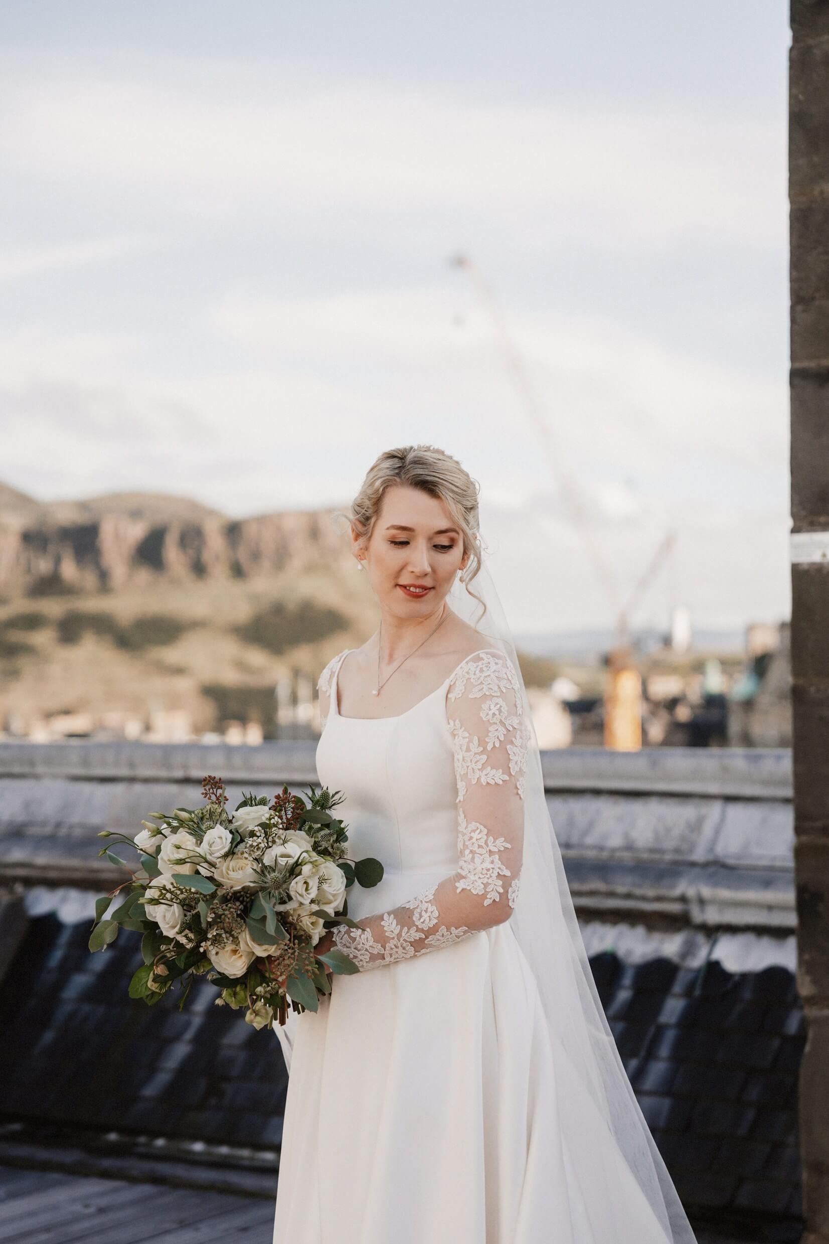 the bride poses for photos on the rooftop of the balmoral hotel edinburgh wedding venue with arthur's seat visible in the background
