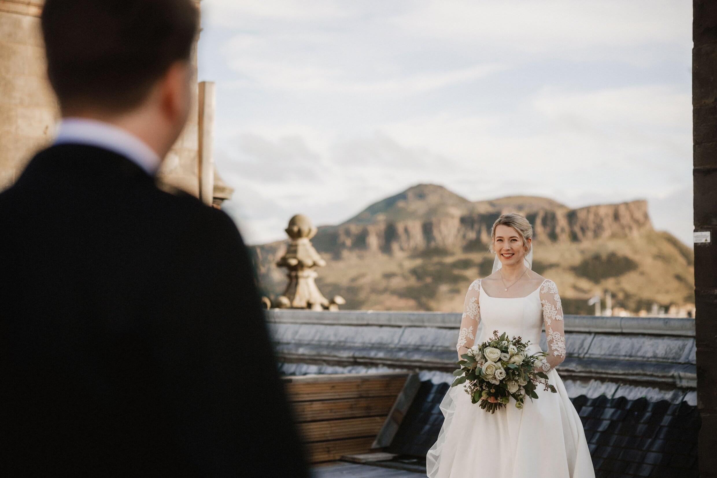 the groom faces the bride on the rooftop of the balmoral hotel edinburgh wedding venue with arthur's seat visible in the background