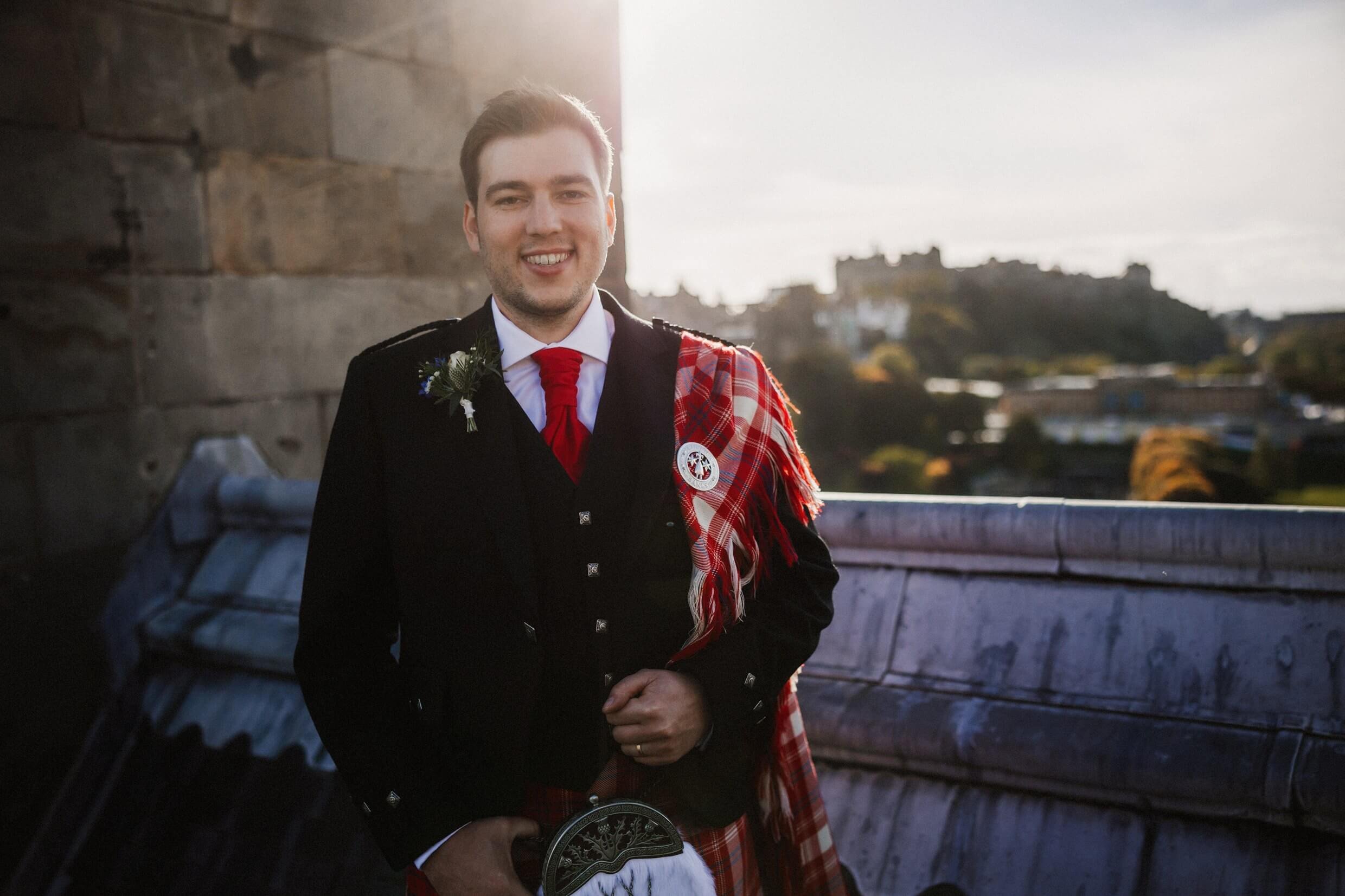 the groom poses for photos on the rooftop of the balmoral hotel edinburgh wedding venue with edinburgh castle visible in the background