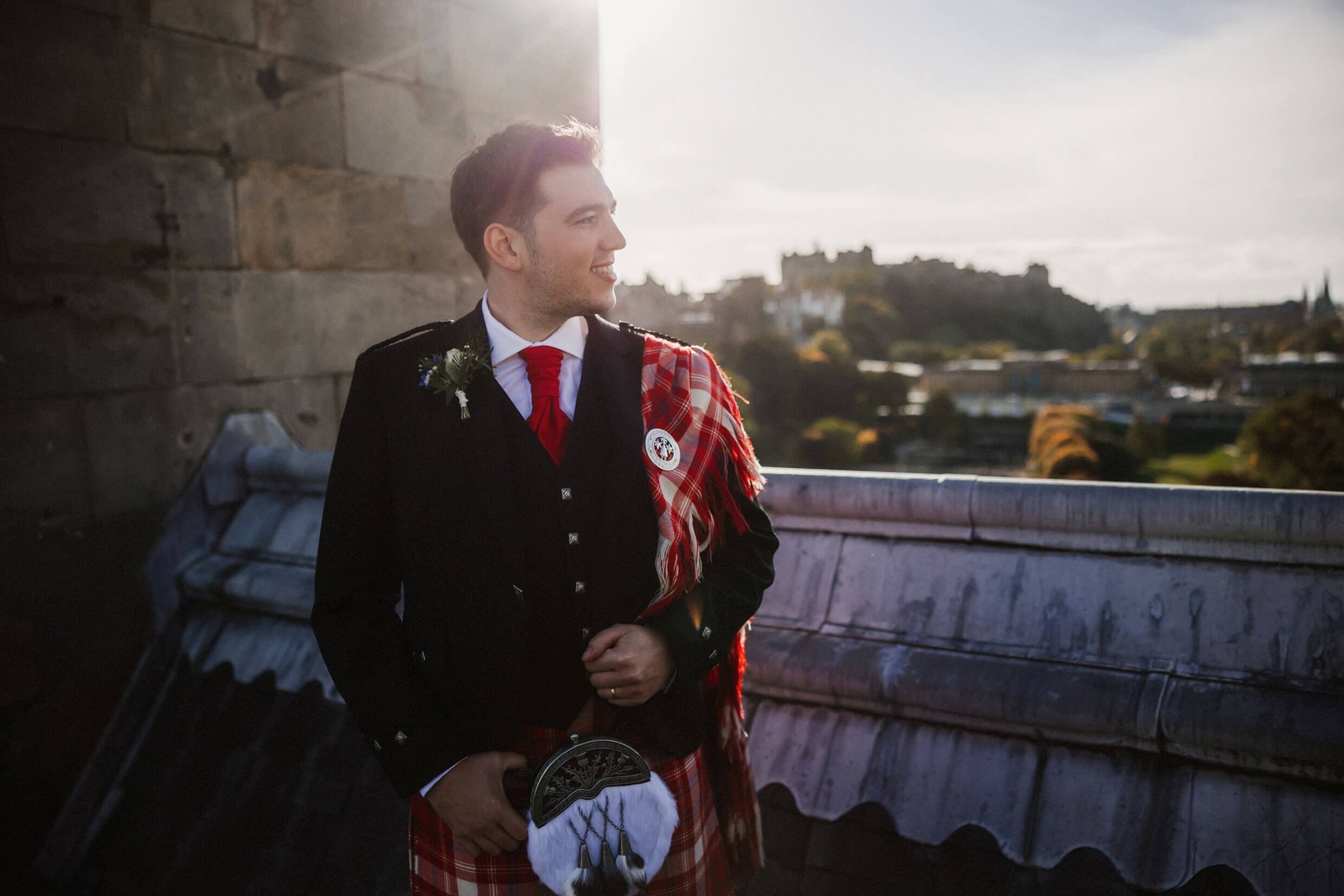 the groom poses for photos on the rooftop of the balmoral hotel edinburgh wedding venue with edinburgh castle visible in the background