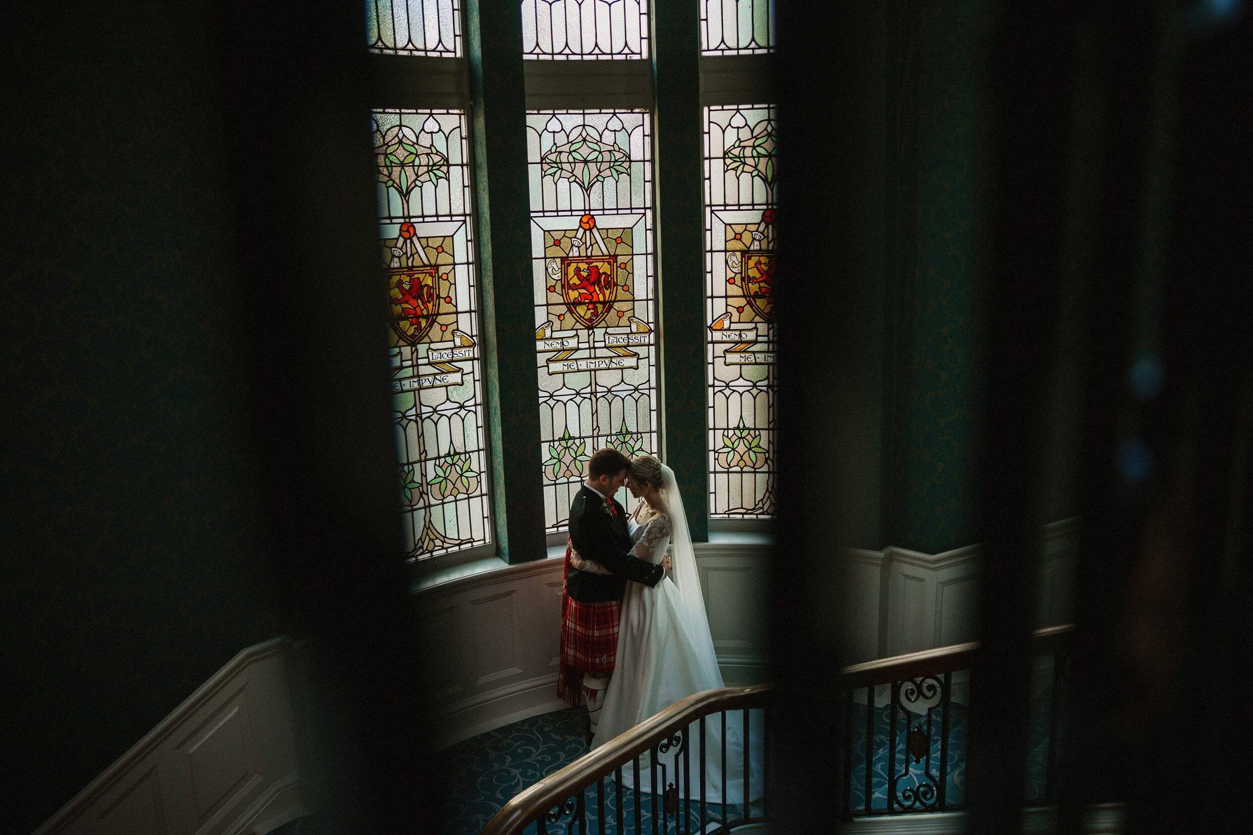 the bride and groom embrace in front of a stained glass window at the balmoral hotel edinburgh wedding venue in scotland