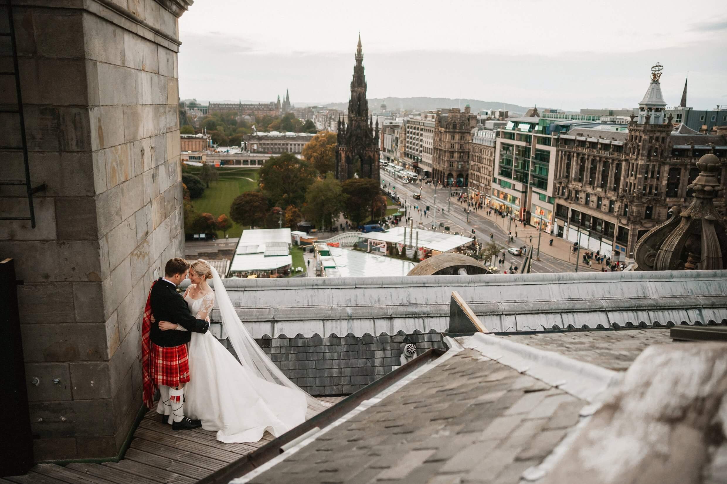 the bride and groom pose for photos on the rooftop of the balmoral hotel edinburgh wedding venue with princes street visible in the background