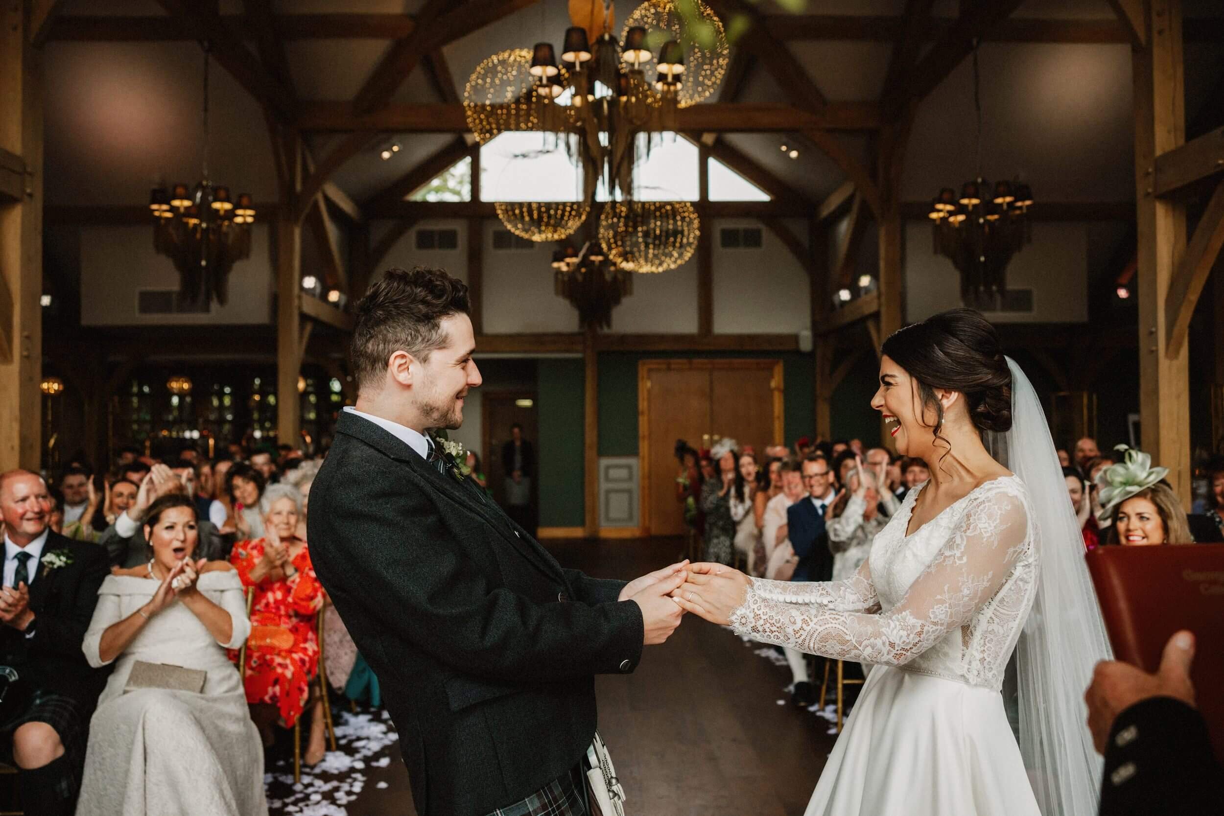 glasgow wedding photographer captures the bride and groom smiling and holding hands at the head of the aisle as guests watch smiling and applauding