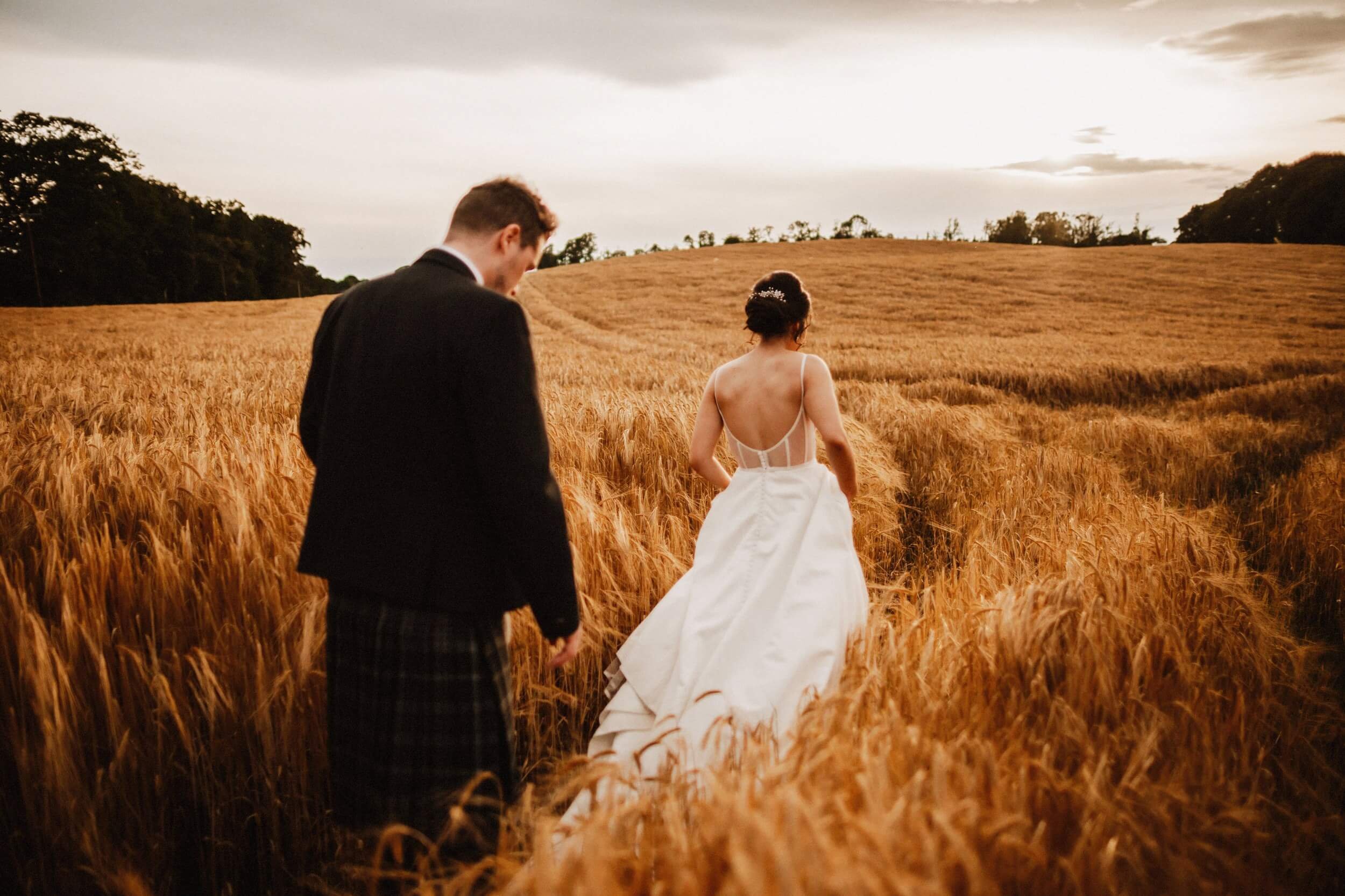 glasgow wedding photographer captures a bride and groom walking through a field of wheat with a cloudy sky above