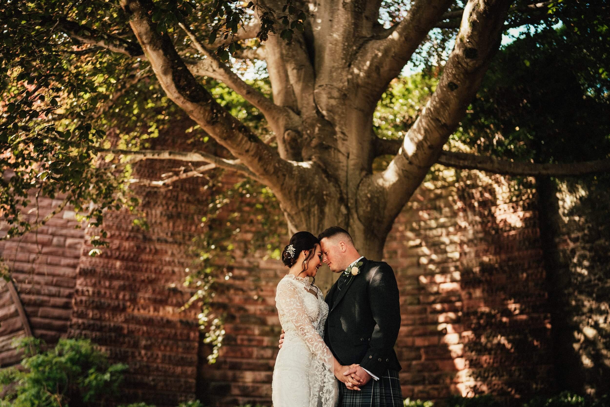 glasgow wedding photographer captures the bride and groom standing forehead to forehead and holding hands in front of a large tree with a red brick wall in the background