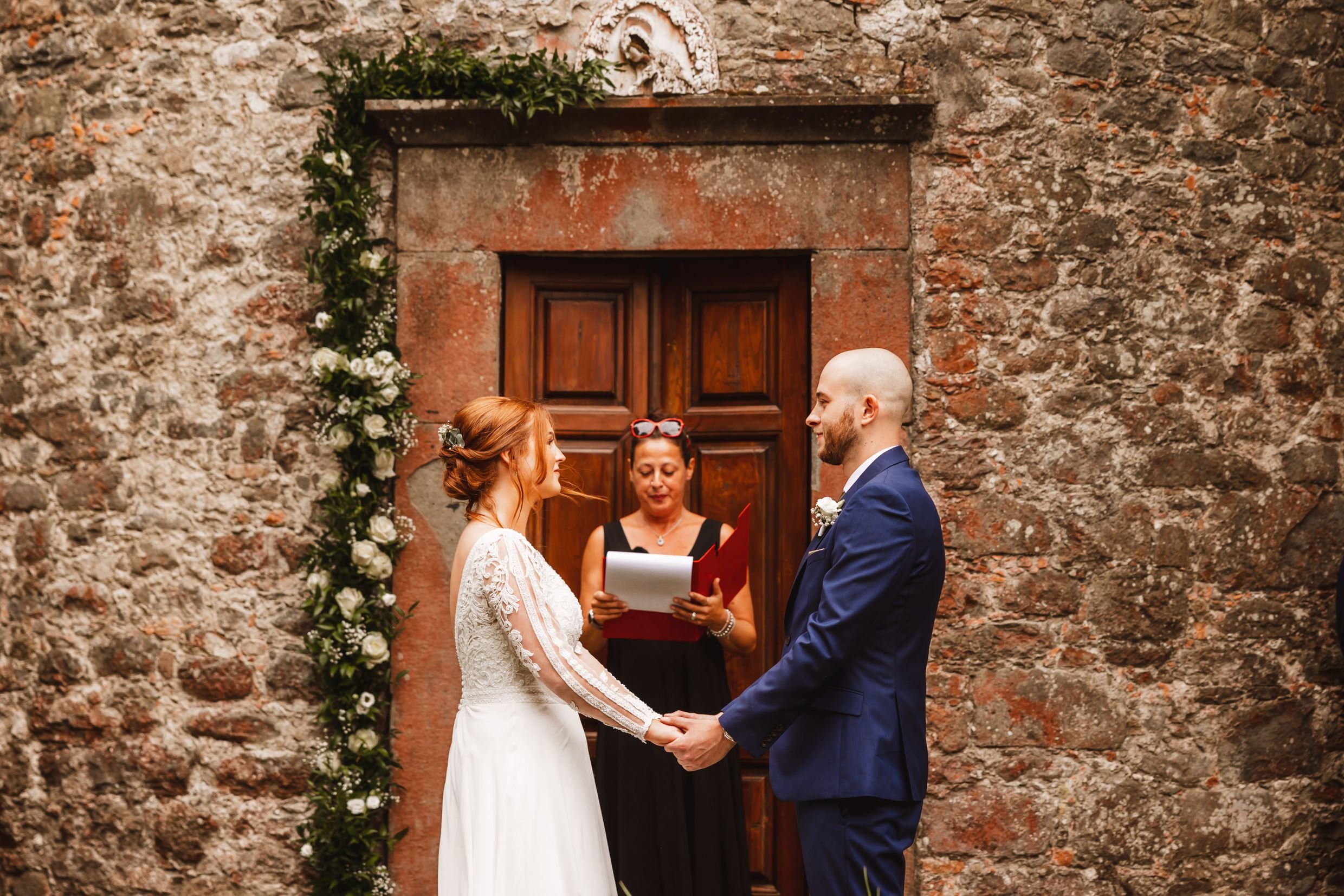 the bride and groom hold hands during wedding ceremony outside the private chapel at the agriturismo la torre italian destination wedding venue in bagni di lucca tuscany