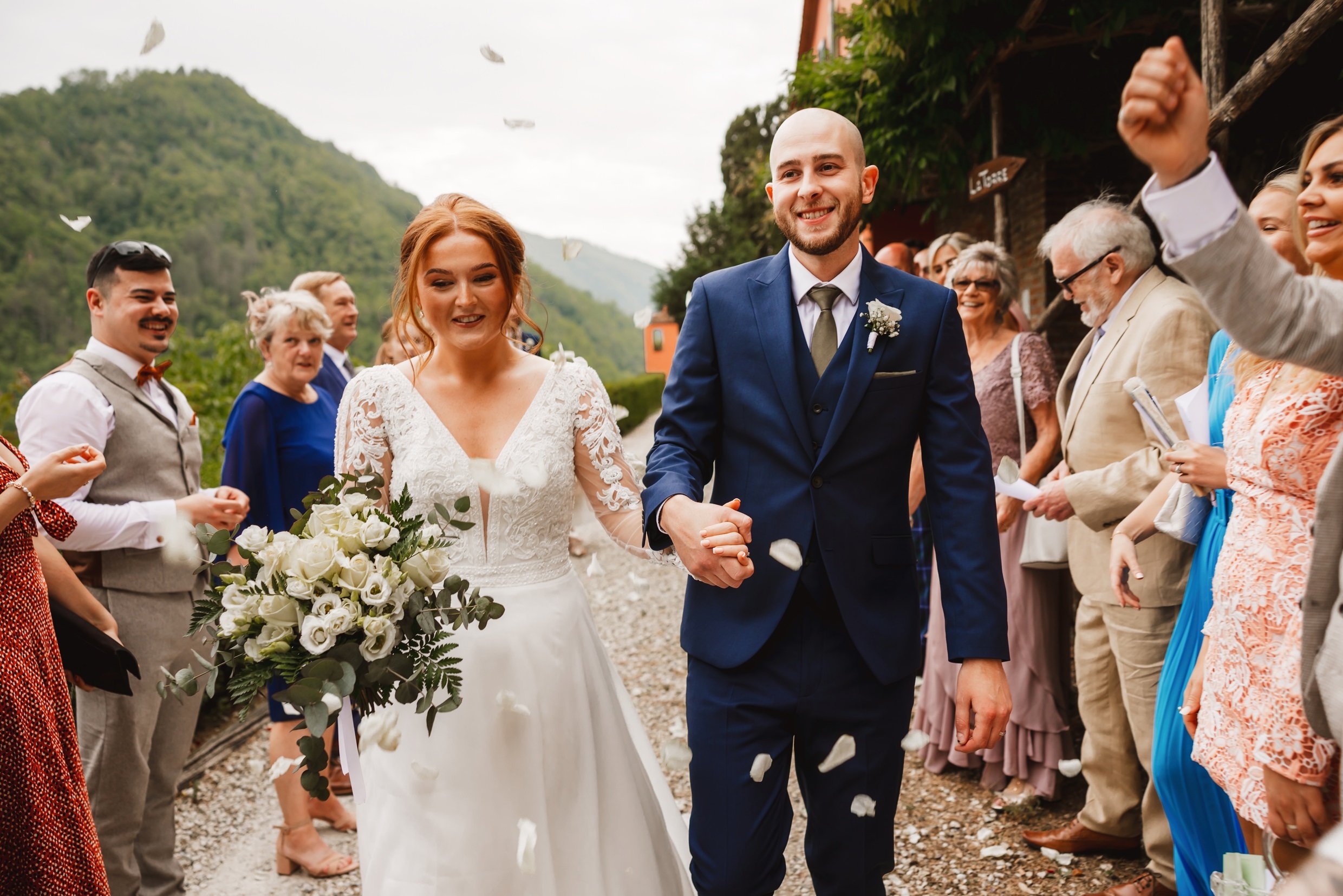 the bride and goom hold hands as wedding guests throw rose petal confetti at the agriturismo la torre italian destination wedding venue in bagni di lucca tuscany