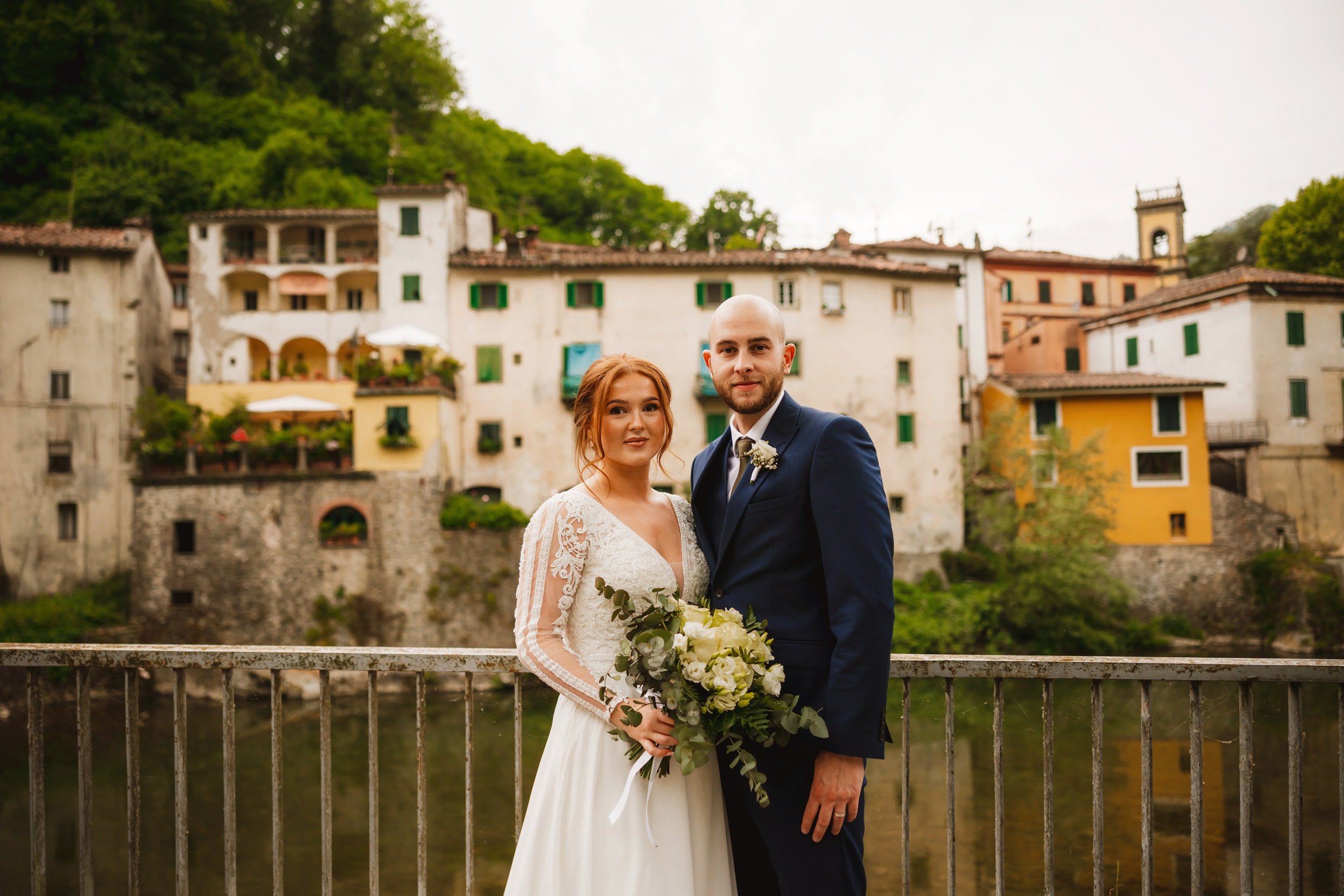 the bride and groom standing by the river lima with old buildings in bagni di luca tuscany in the background following their italian destination wedding