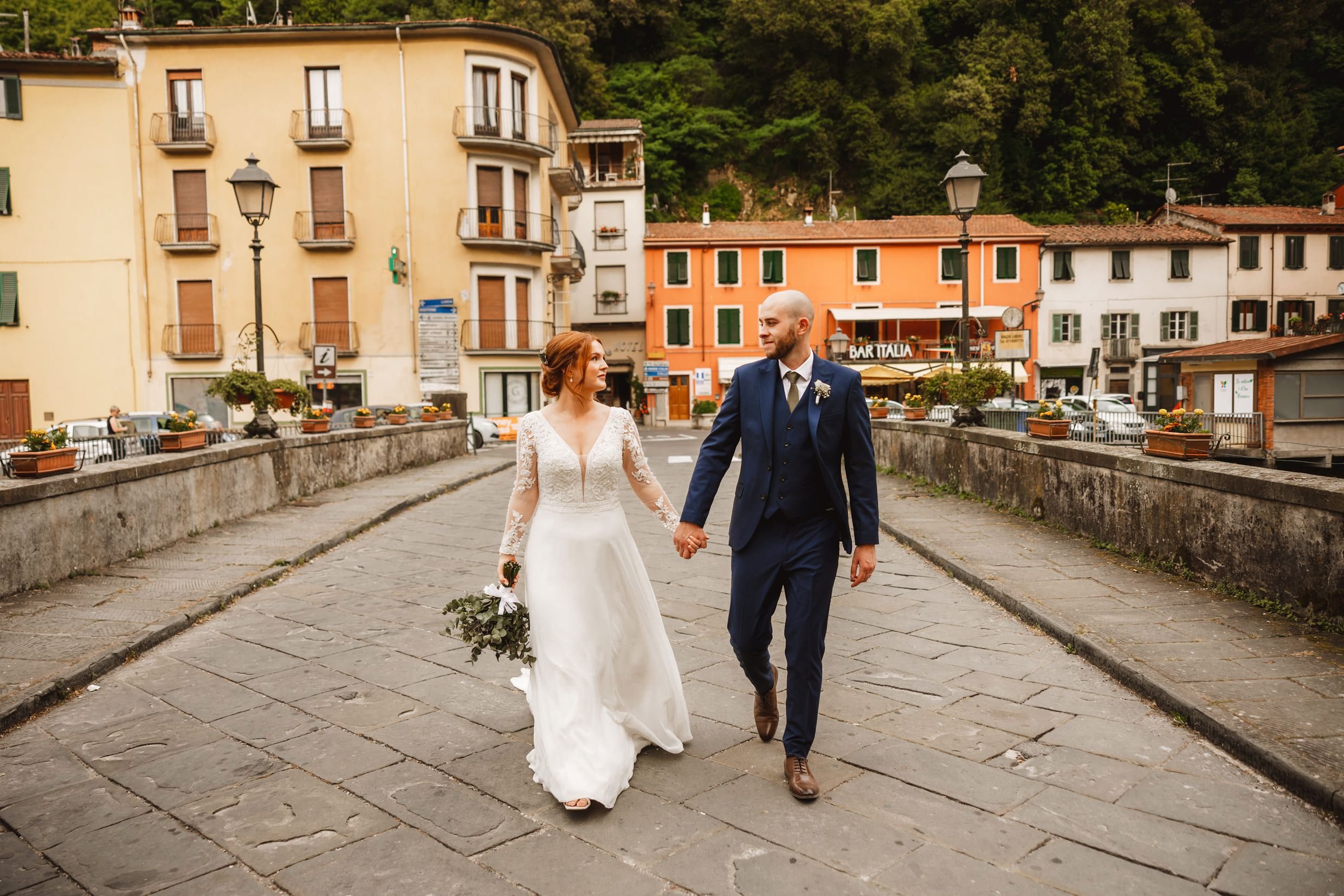 the bride and groom hold hands while walking across a bridge in bagni di lucca in tuscany during their italian destination wedding