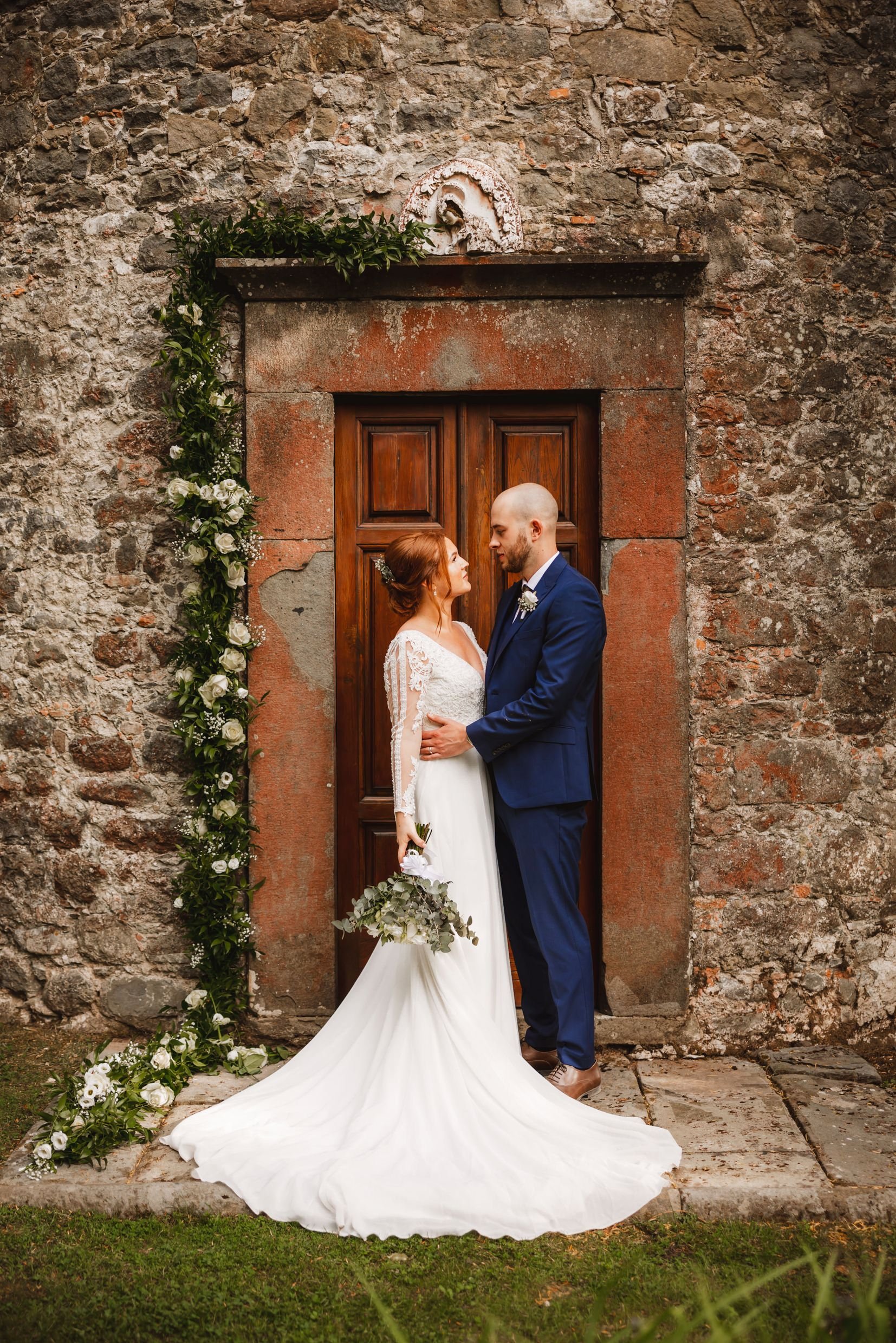 the bride and groom standing in front of a wooden door in an old stone building in bagni di lucca in tuscany during their italian destination wedding