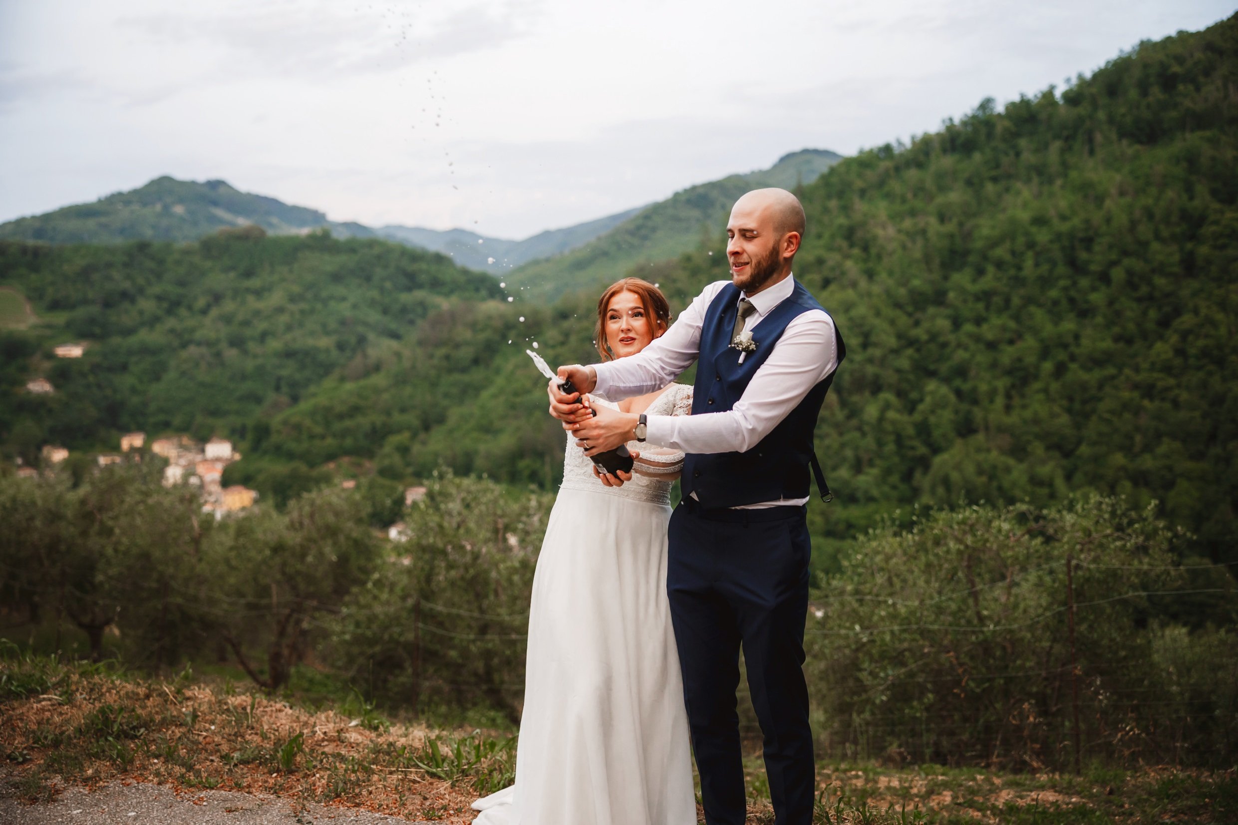 the bride and groom open a champagne bottle with hills and trees in the background at the agriturismo la torre italian destination wedding venue in bagni di lucca tuscany