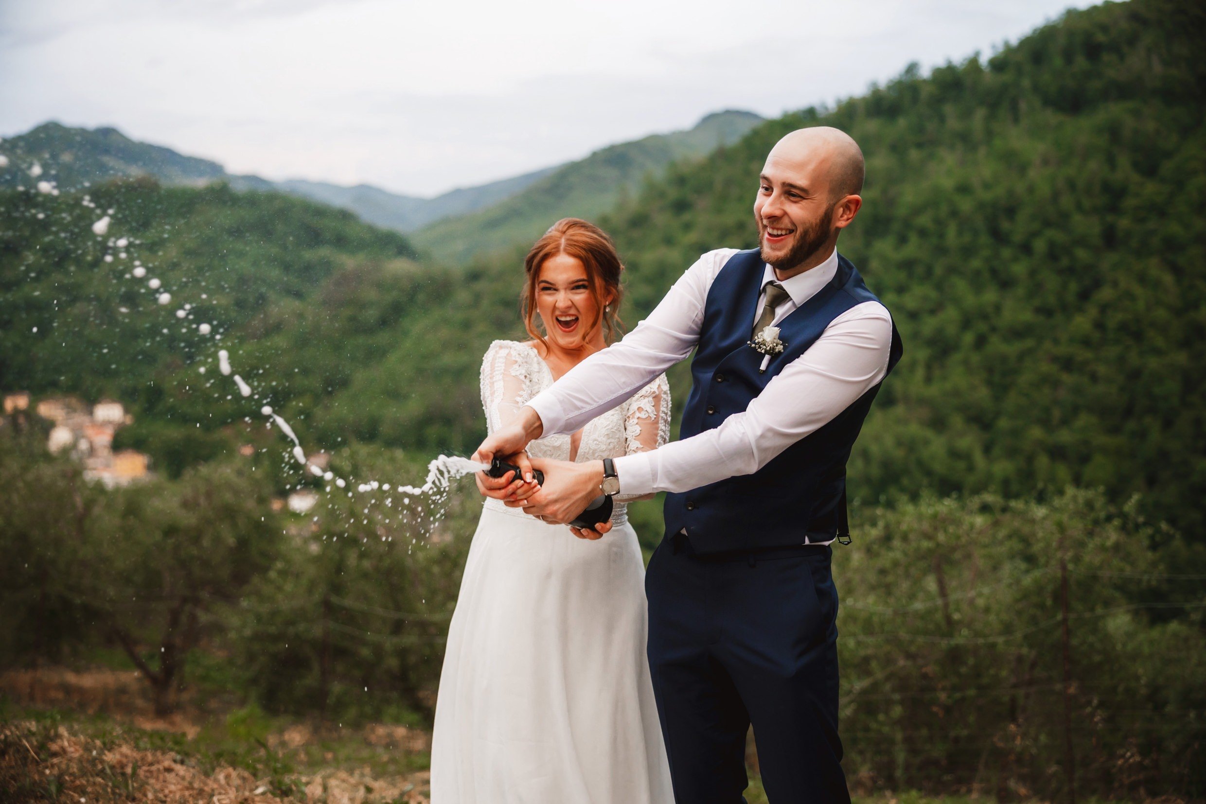 the bride and groom spray champagne with hills and trees in the background at the agriturismo la torre italian destination wedding venue in bagni di lucca tuscany