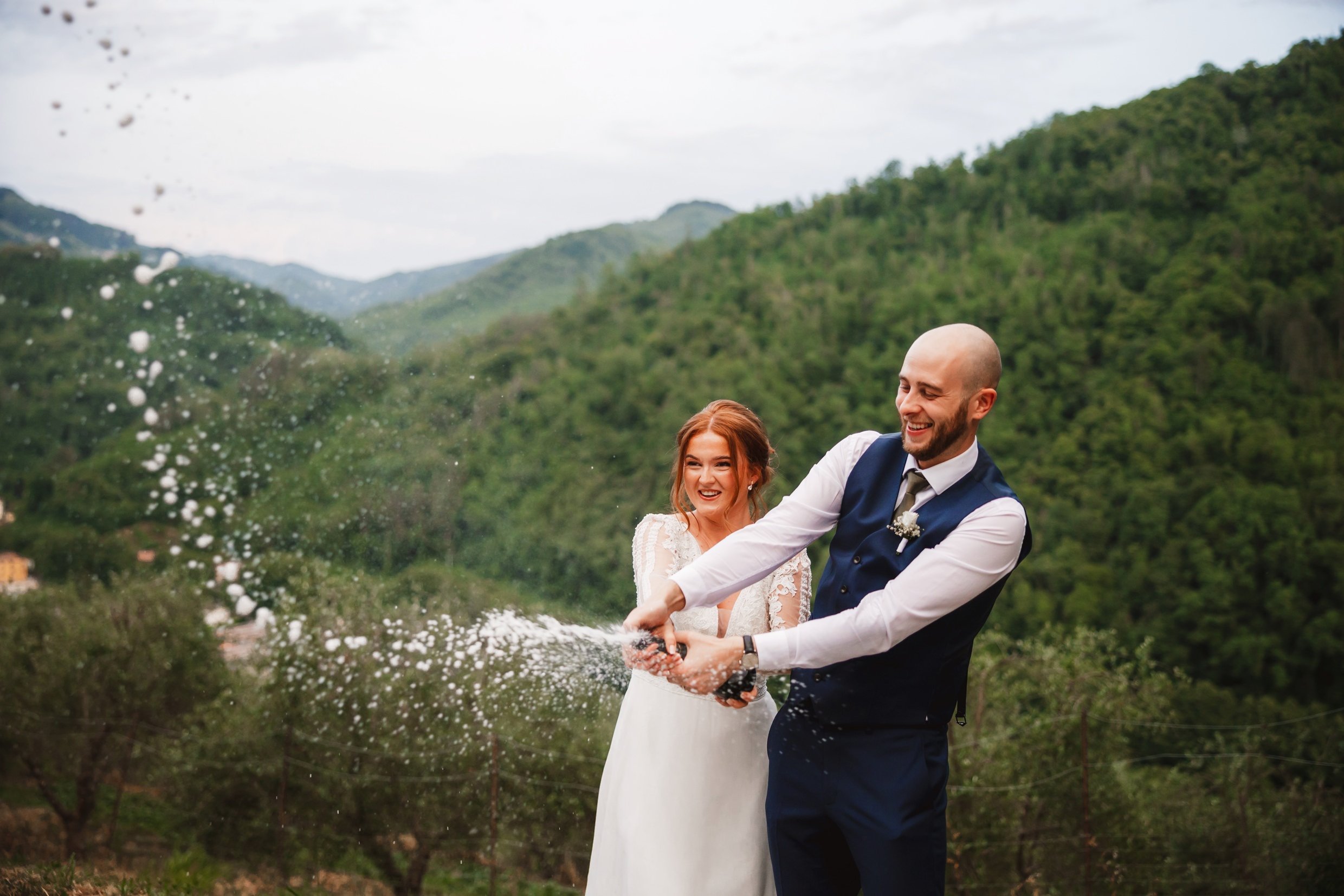 the bride and groom spray champagne with hills and trees in the background at the agriturismo la torre italian destination wedding venue in bagni di lucca tuscany