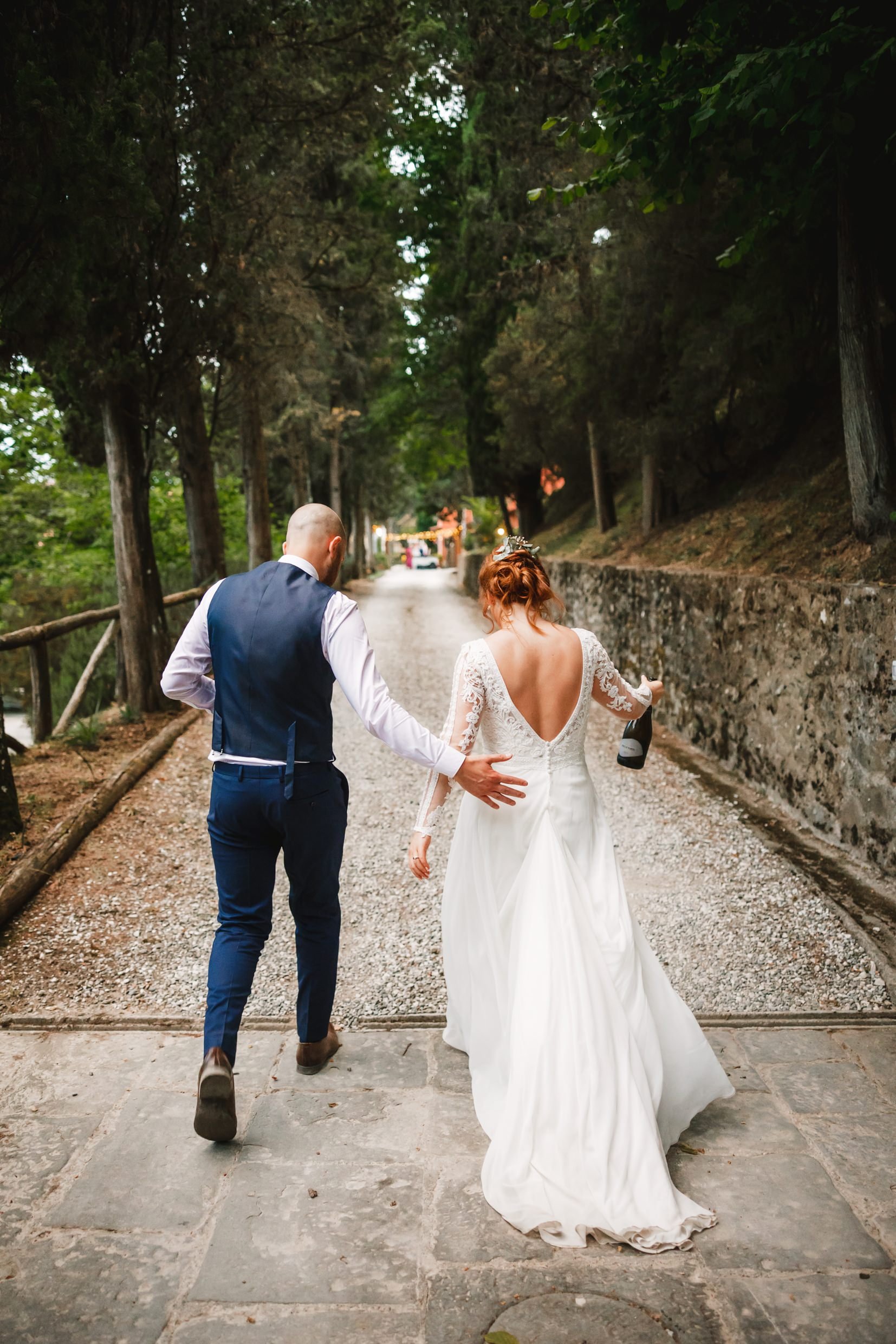 the bride and groom walk along a path beneath trees towards the agriturismo la torre italian destination wedding venue in bagni di lucca tuscany