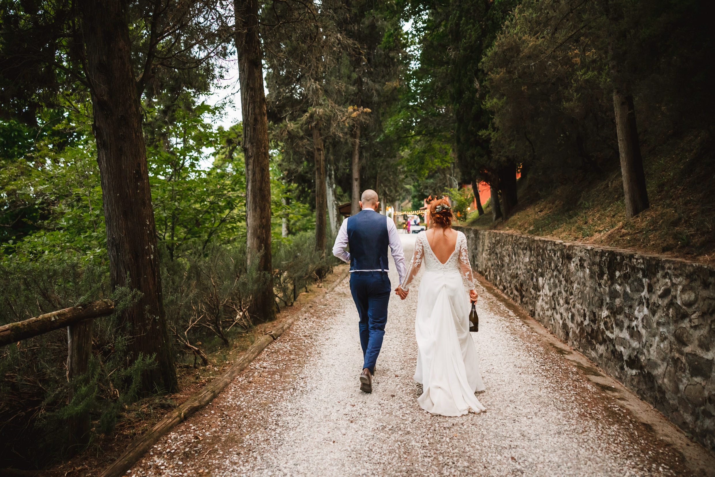 the bride and groom walk along a path beneath trees towards the agriturismo la torre italian destination wedding venue in bagni di lucca tuscany