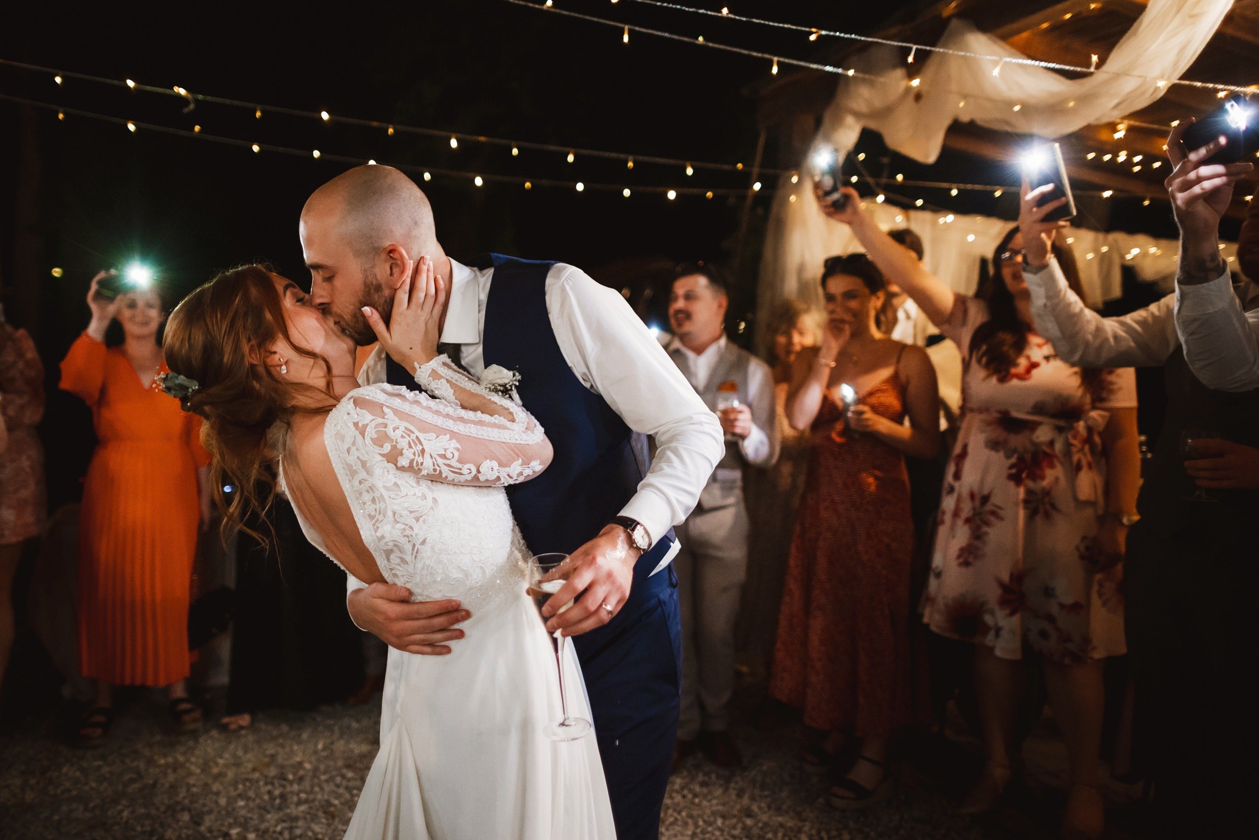 the bride and groom kiss underneath fairy lights as guests look on outside at the agriturismo la torre italian destination wedding venue in bagni di lucca tuscany