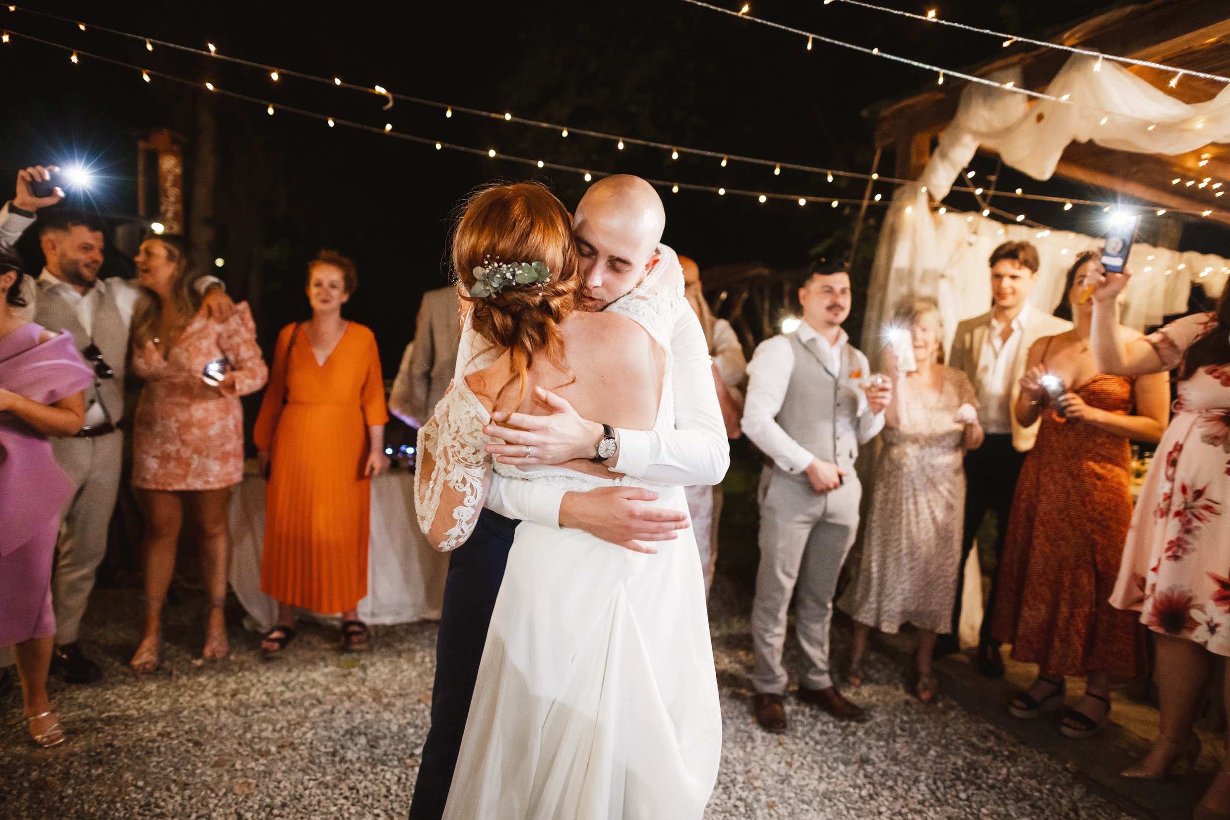 the bride and groom hug underneath fairy lights as guests look on outside at the agriturismo la torre italian destination wedding venue in bagni di lucca tuscany