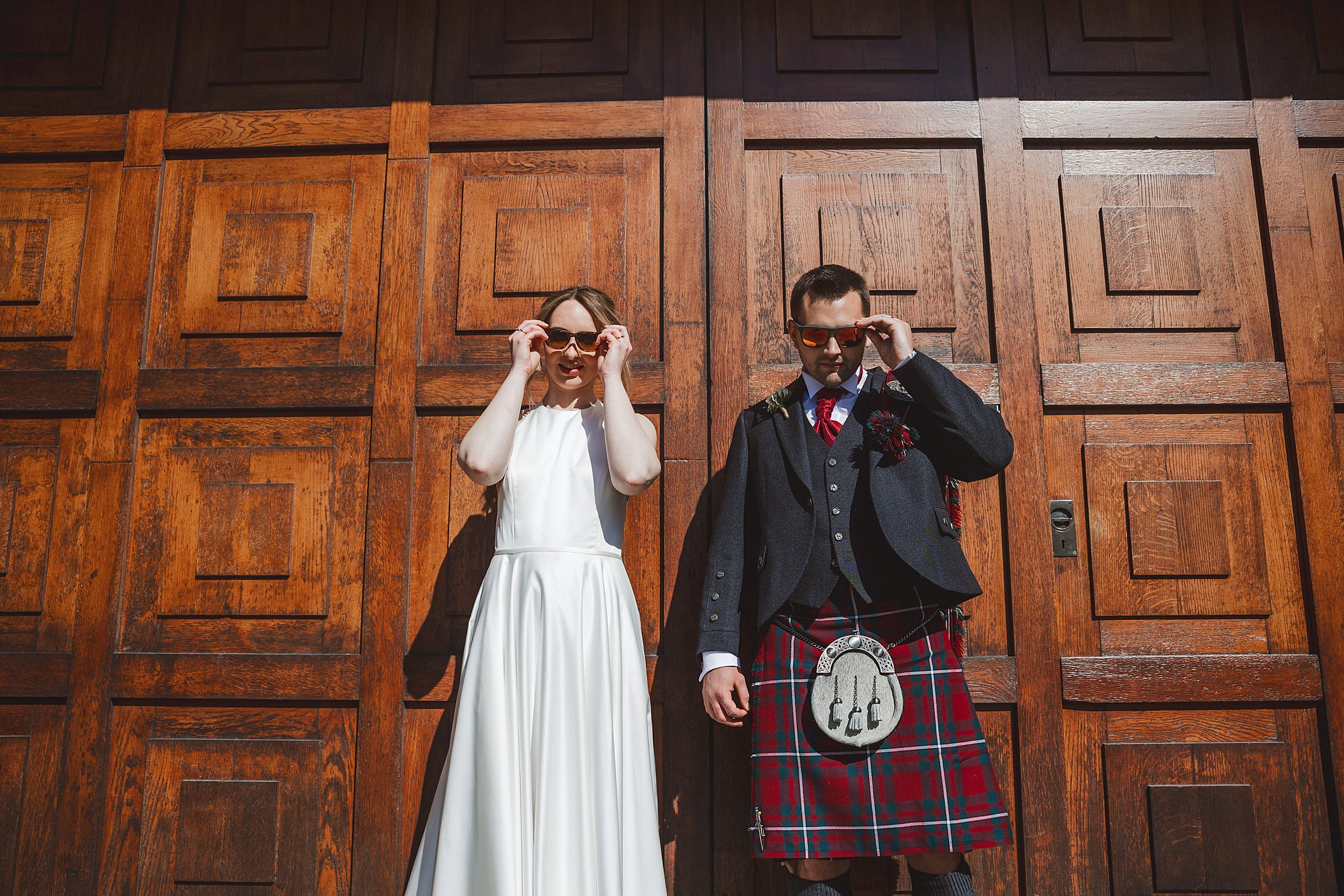 editorial shot of the bride and groom wearing sunglasses and holding hands in front of a wooden door by documentary wedding photographer edinburgh scotland