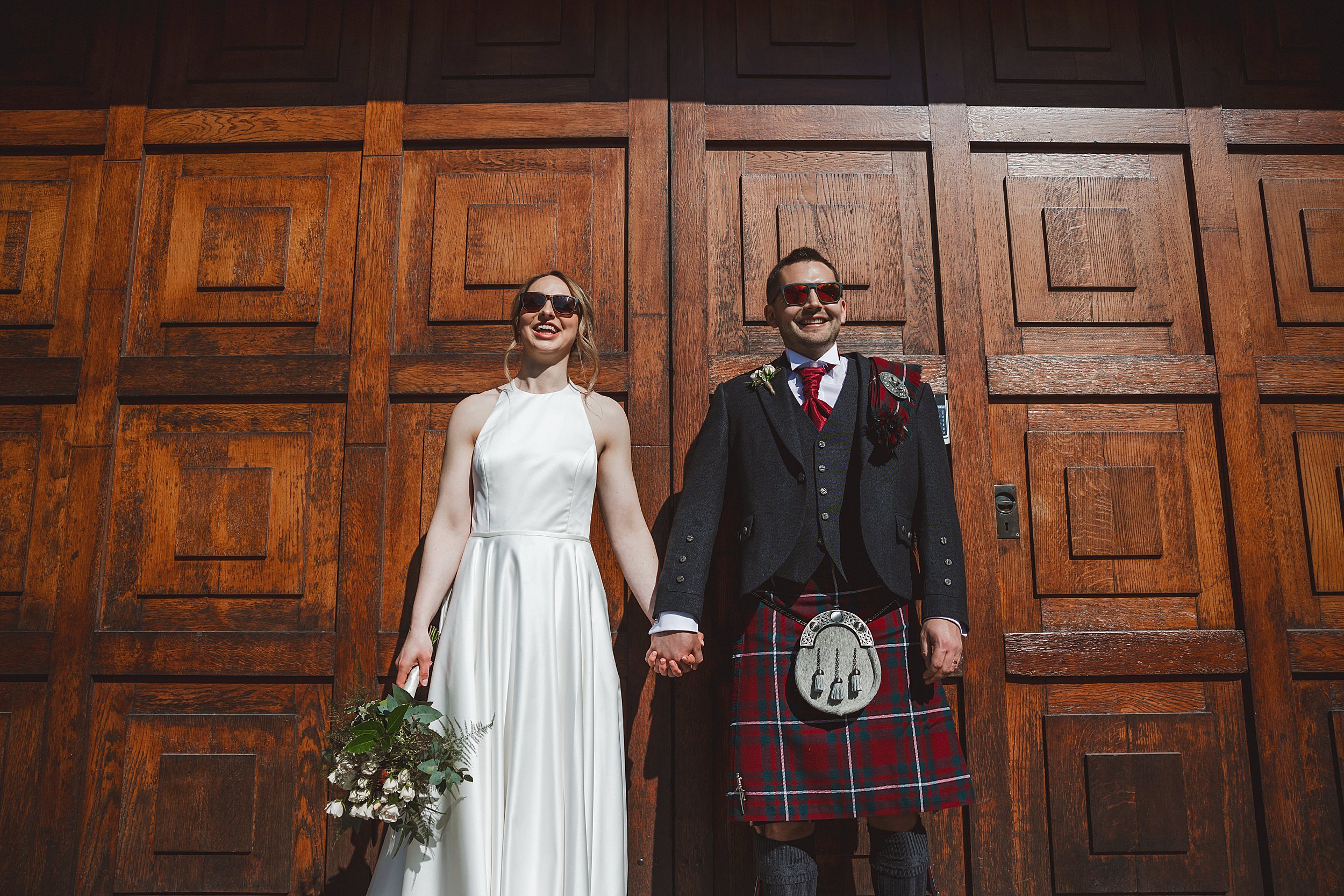 editorial shot of the bride and groom wearing sunglasses and holding hands in front of a wooden door by documentary wedding photographer edinburgh scotland