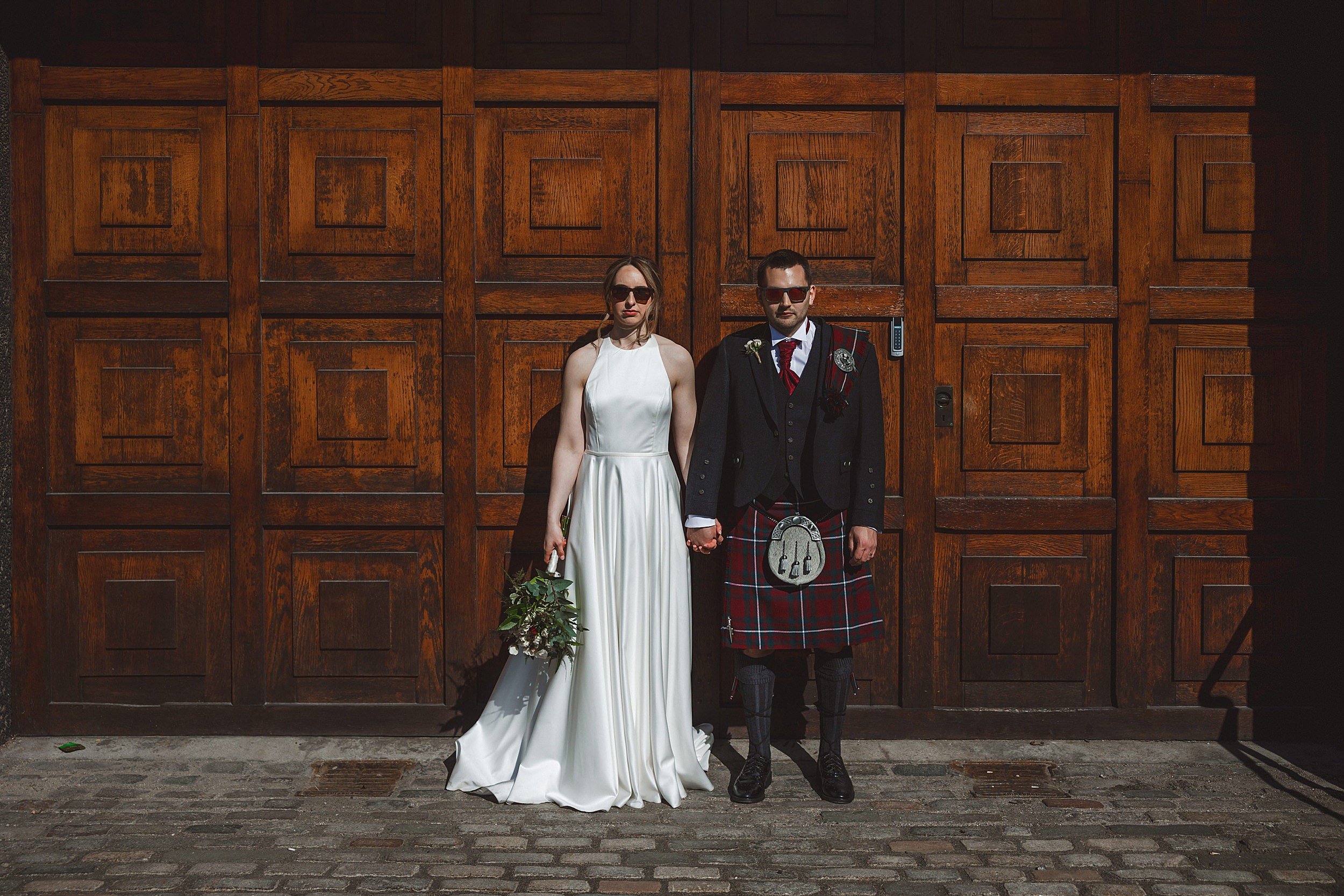 editorial shot of the bride and groom wearing sunglasses and holding hands in front of a wooden door by documentary wedding photographer edinburgh scotland