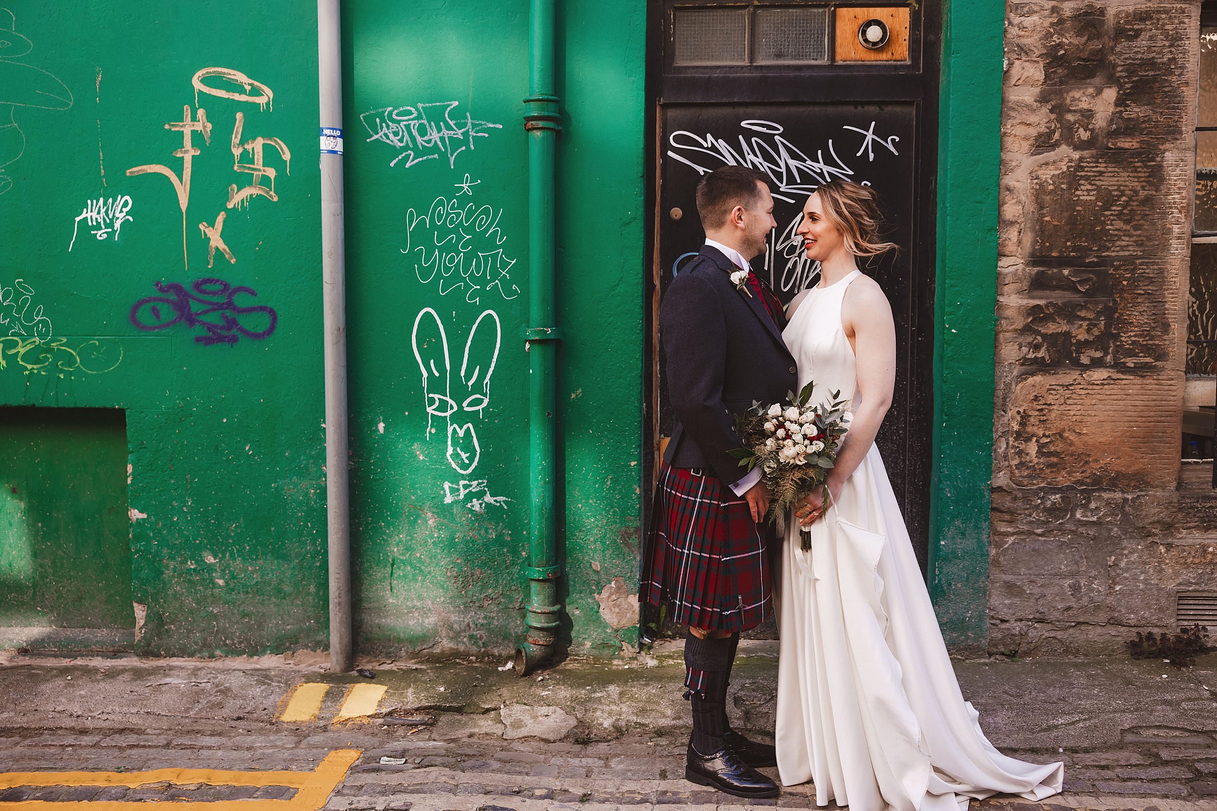 editorial shot of the bride and groom facing each other standing in an edinburgh street in front of a graffitied wall by documentary wedding photographer edinburgh
