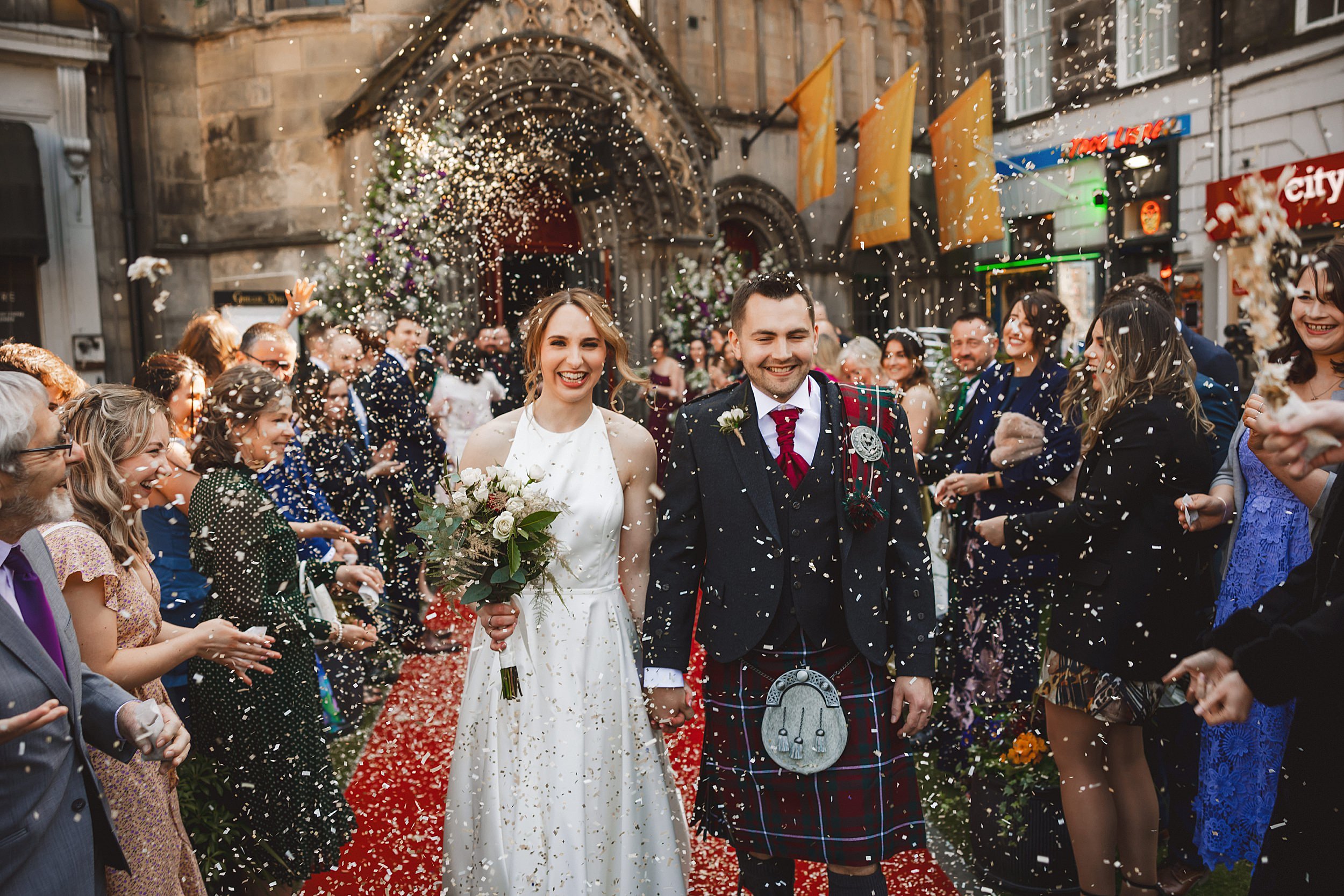reportage shot of the bride and groom leaving their edinburgh wedding venue holding hands while the wedding party throw confetti by documentary wedding photographer edinburgh