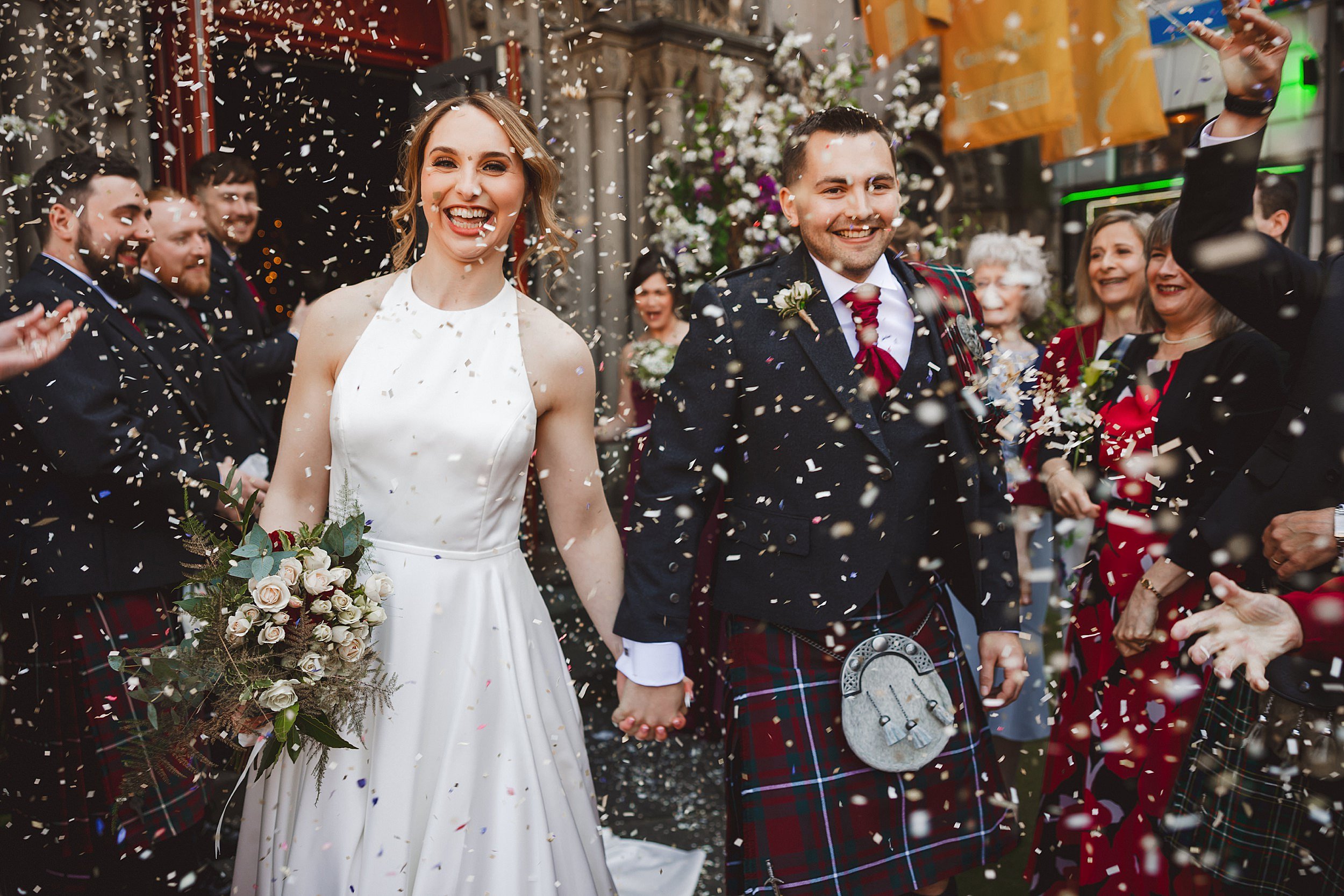 reportage shot of the bride and groom leaving their edinburgh wedding venue holding hands while the wedding party throw confetti by documentary wedding photographer edinburgh