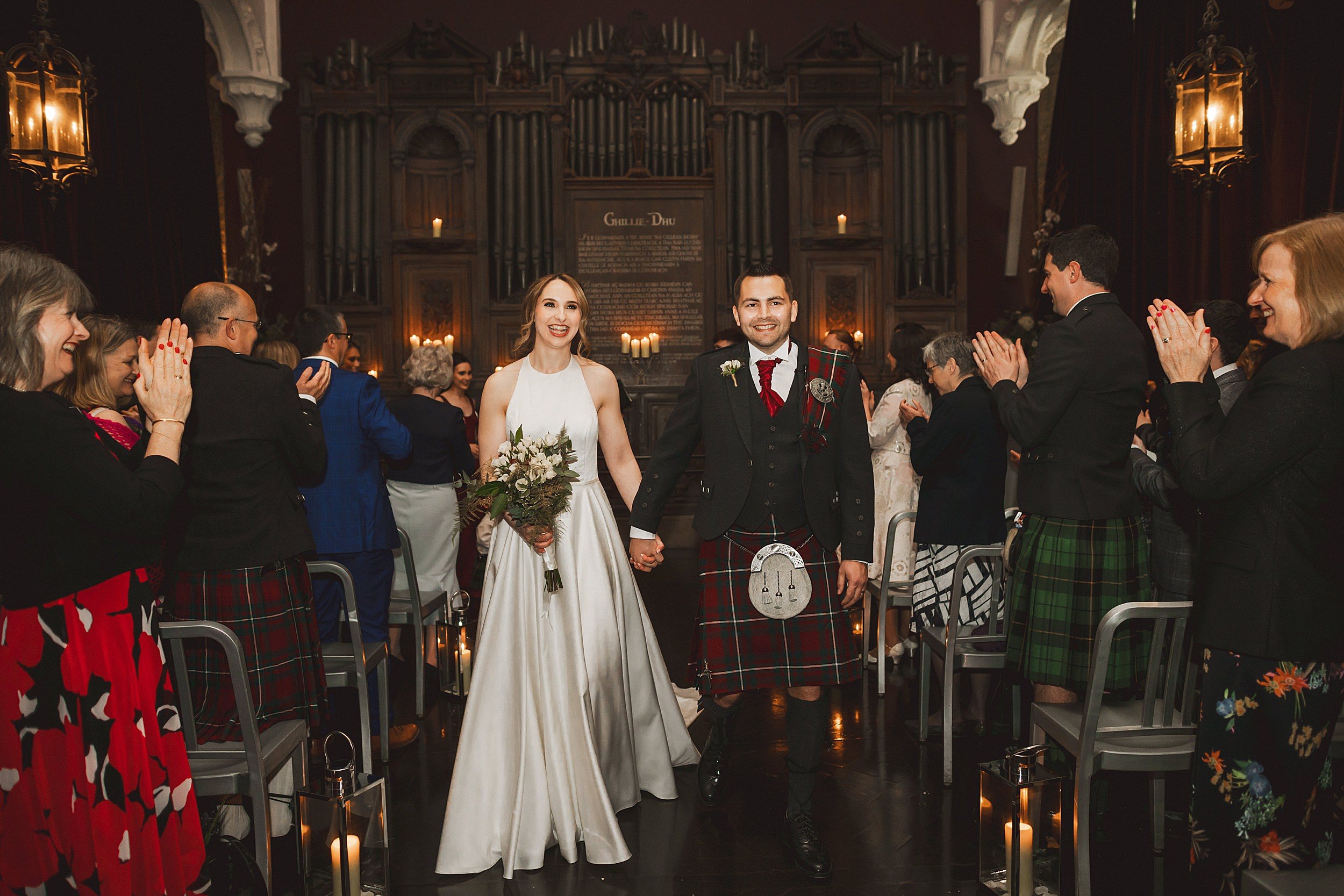 guests stand and applaud as the bride and groom walk down the aisle together holding hands at the ghillie dhu edinburgh wedding venue in scotland shot by documentary wedding photographer edinburgh