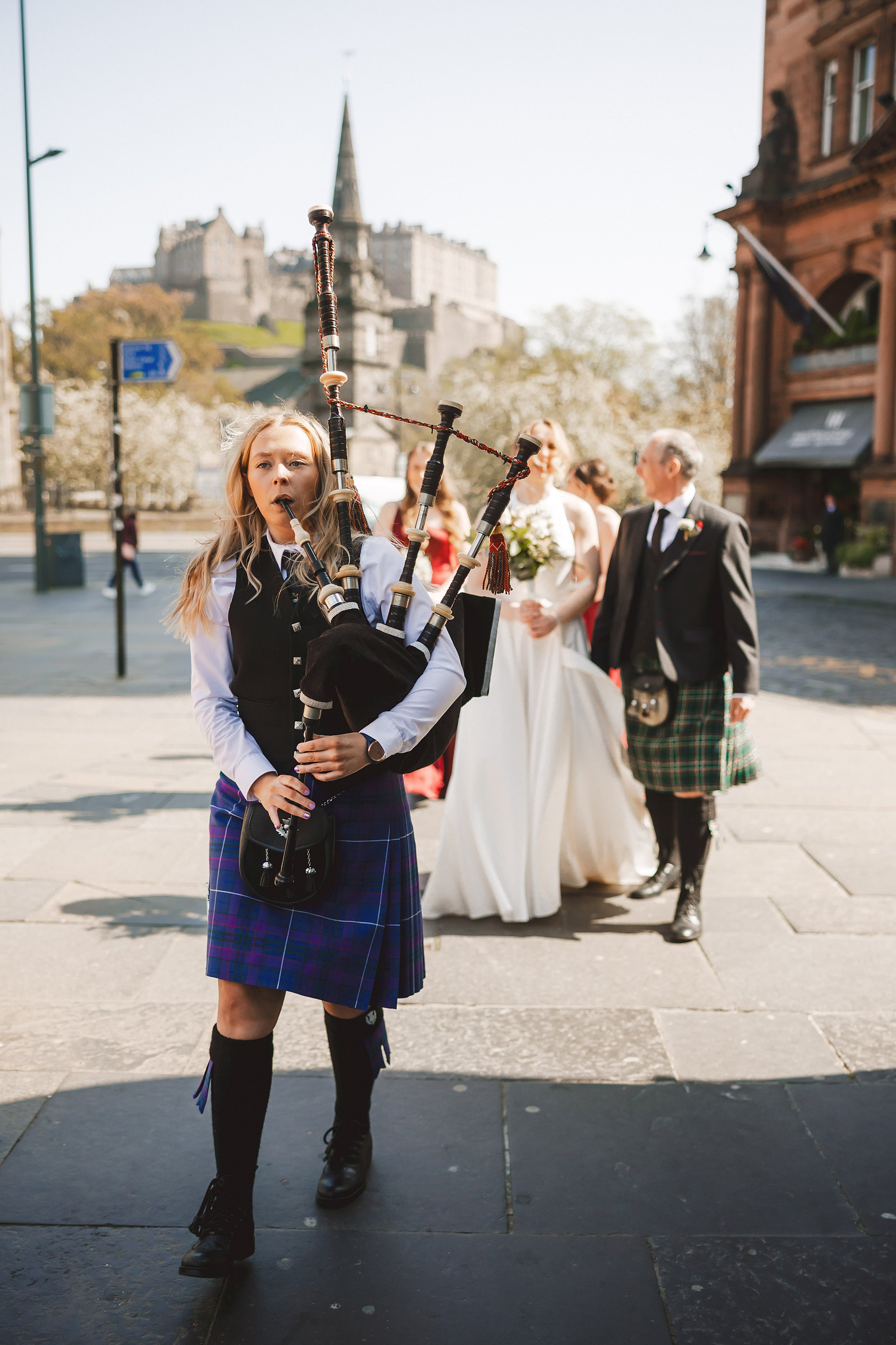 reportage shot of a female piper leading the bridal party to the ghillie dhu with edinburgh castle visible in the background by documentary wedding photographer edinburgh