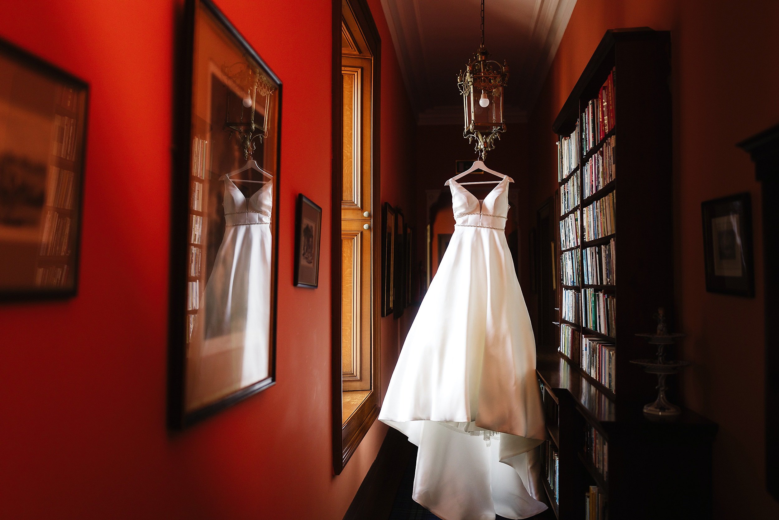 wedding dress hanging in front of a bookcase in a red walled passageway in errol park wedding venue in perthshire scotland