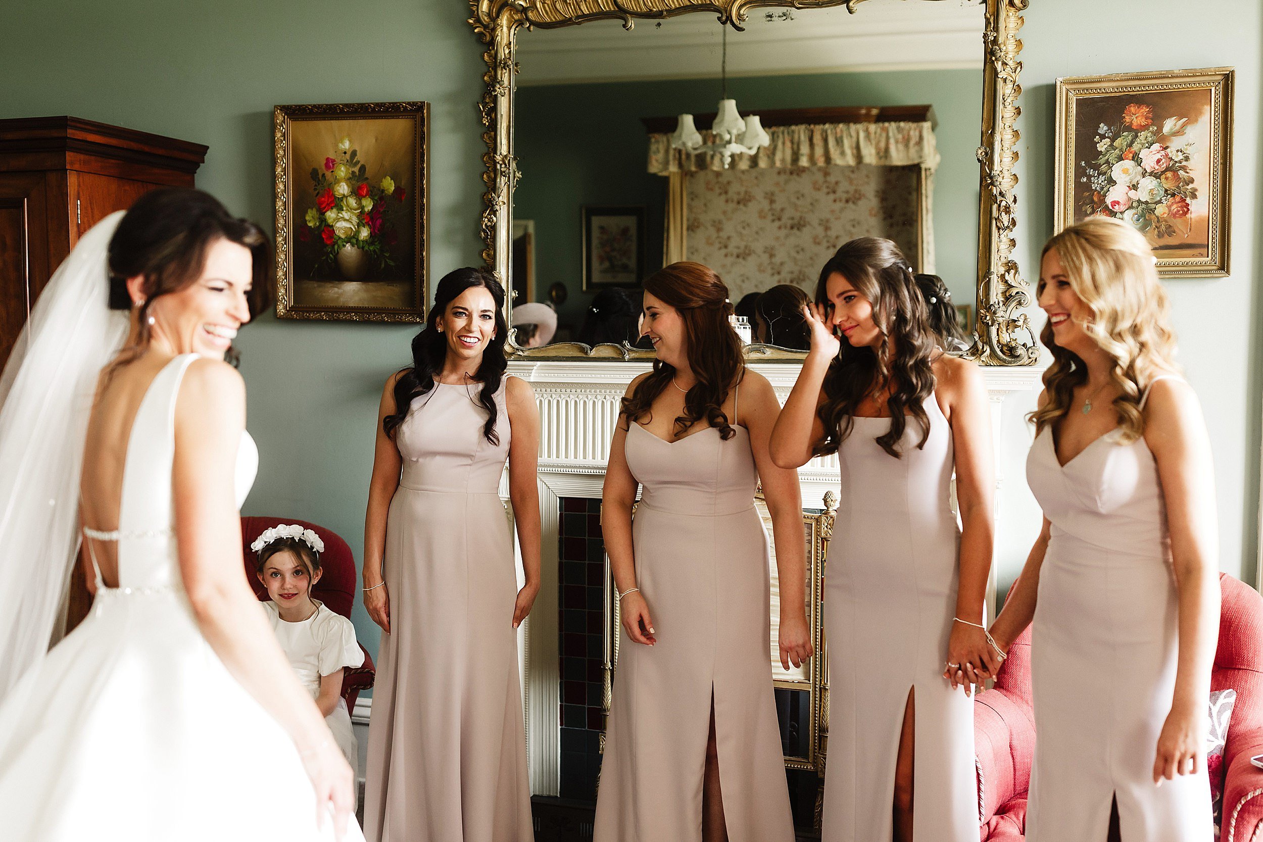 four bridesmaids wearing long blush pink gowns greet the bride in front of an ornate gold mirror above a fireplace in a bedroom in errol park wedding venue in perthshire scotland
