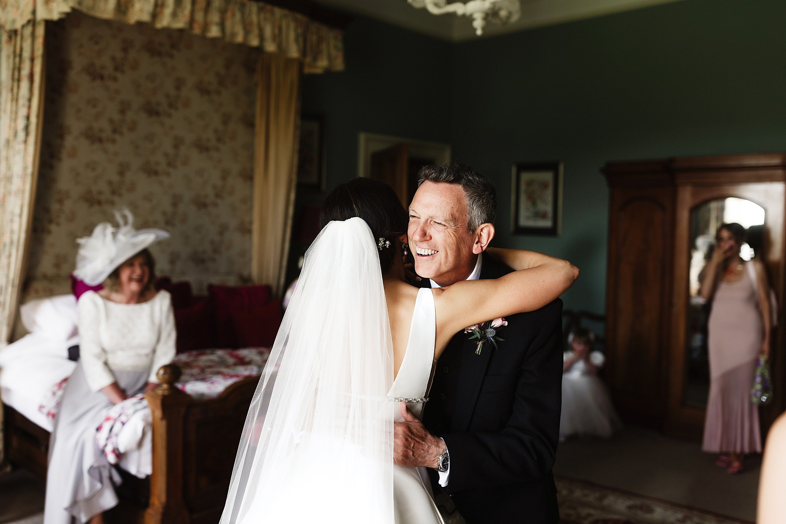 the bride hugs her father as the mother of the bride and a bridesmaid look on in a bedroom in errol park wedding venue in perthsire scotland