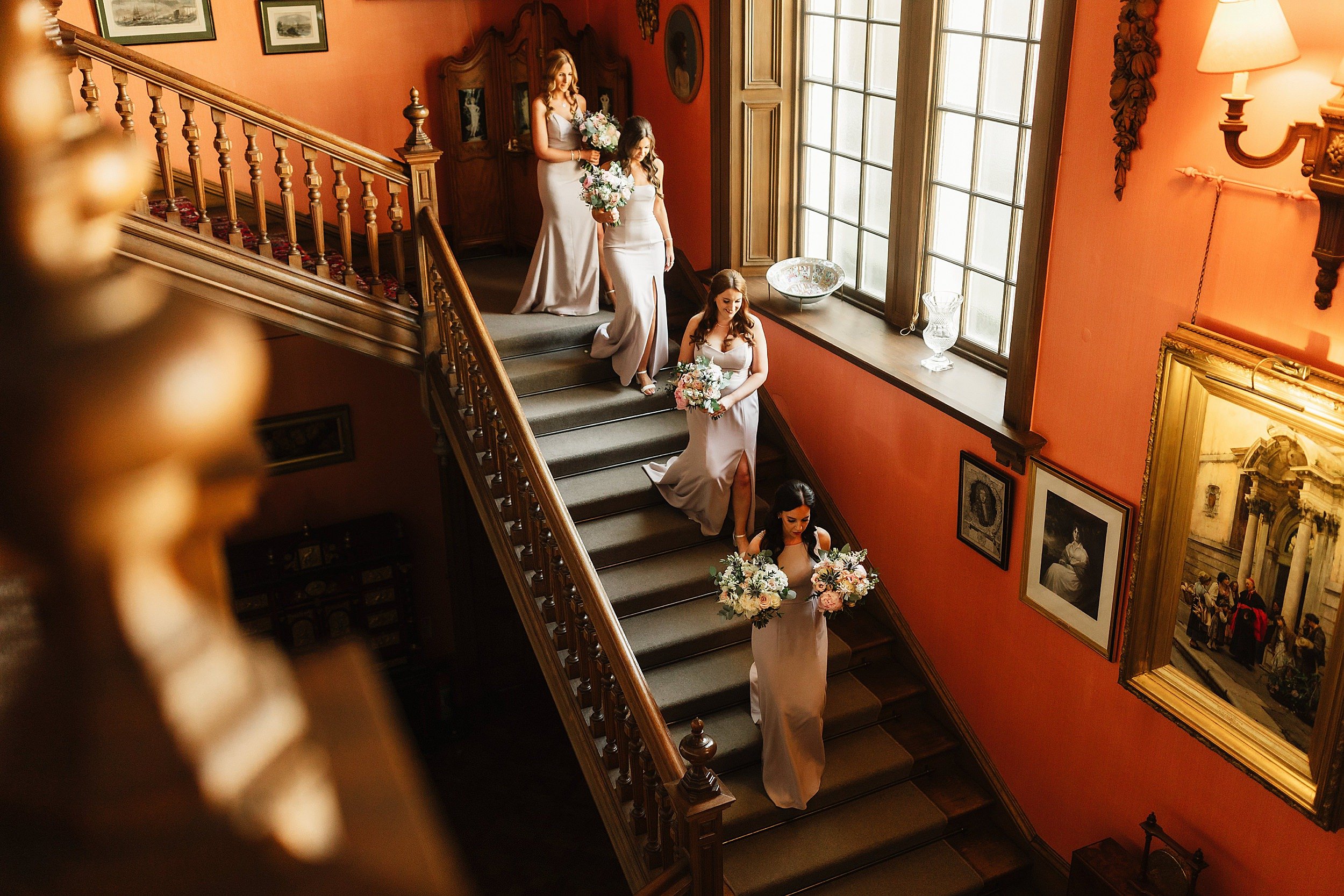 four bridesmaids wearing blush pink gowns and carrying bouquets descend a staircase in errol park wedding venue in perthshire in scotland