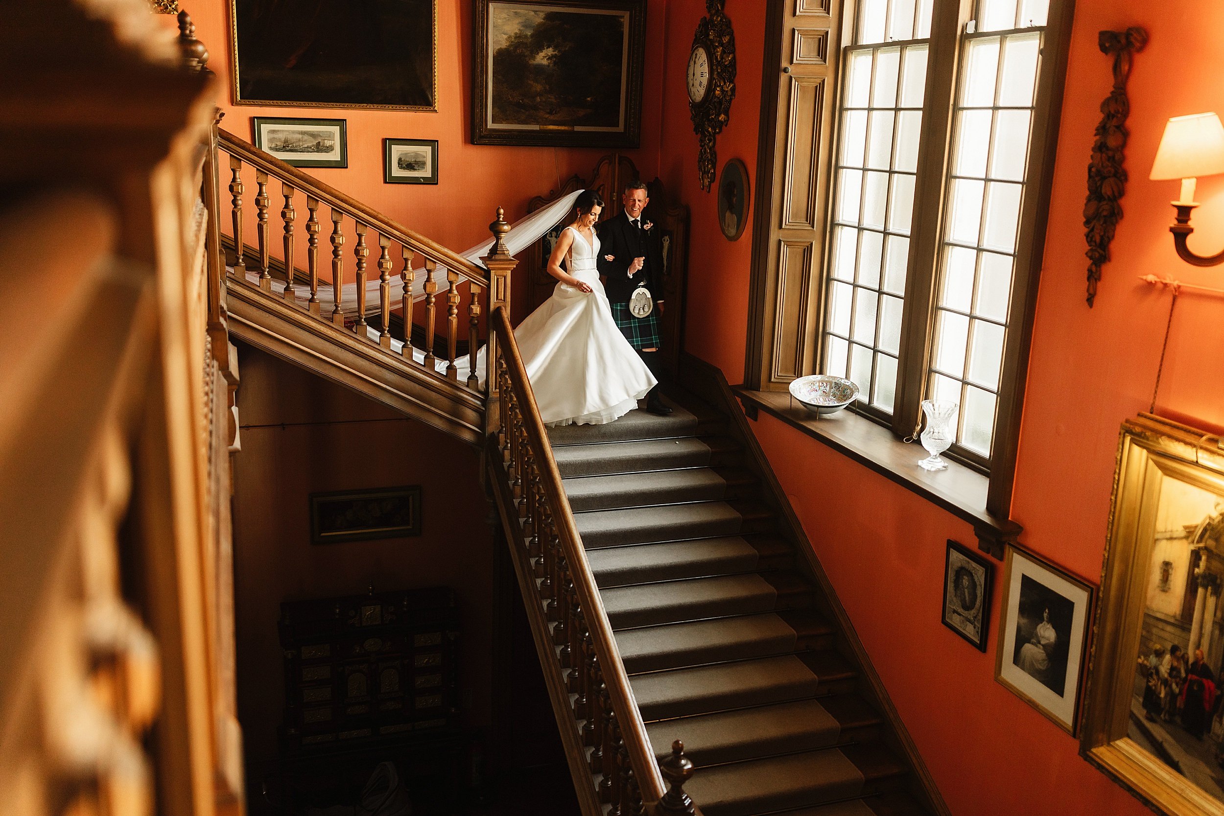 the bride wearing a long white gown and veil and the father of the bride descend a staircase in errol park wedding venue in perthshire in scotland
