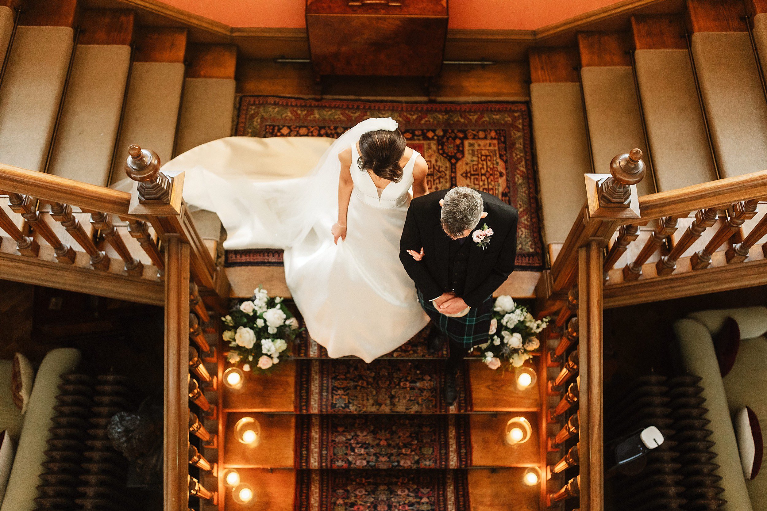 view from above of the bride wearing a long white gown and veil and the father of the bride descending a staircase in errol park wedding venue in perthshire in scotland