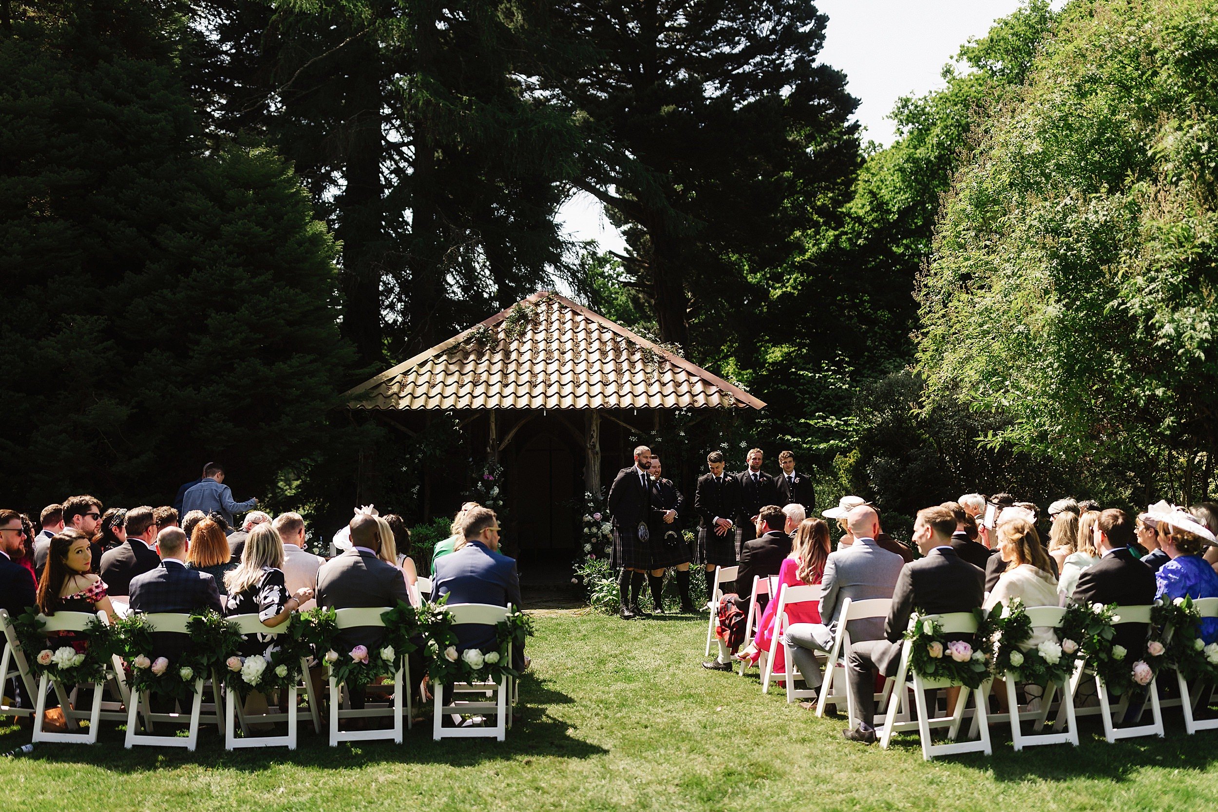 wedding guests are seated on white chairs in front of a wooden gazebo in the gardens of errol park wedding venue in perthshire scotland
