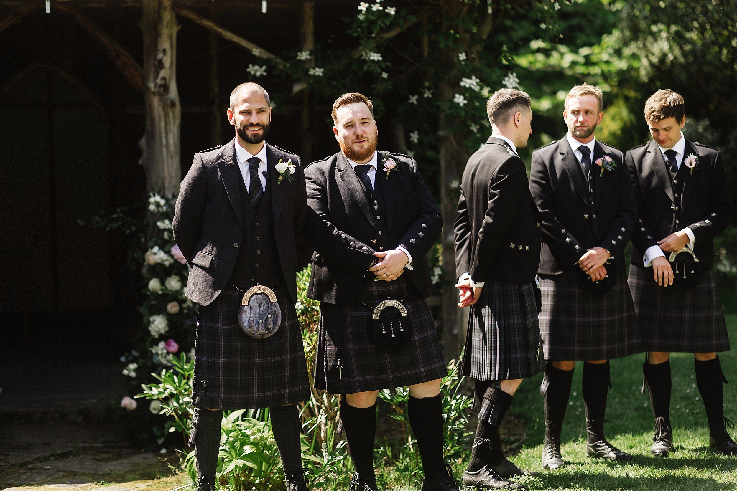 the groom and groomsmen wearing kilts and traditional scottish attire stand in front of a wooden gazebo in the grounds of errol park wedding venue in perthshire in scotland