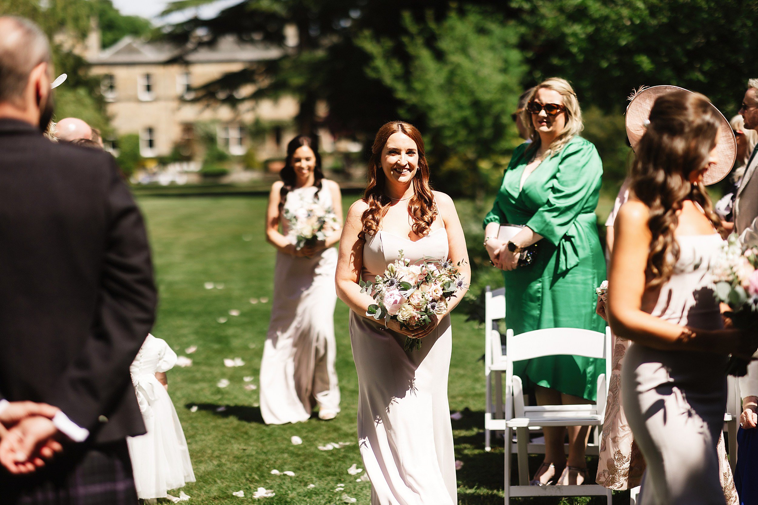 guests look on as two bridesmaids wearing long blush pink gowns and carrying bouquets walk down the aisle in the gardens of errol park wedding venue in scotland