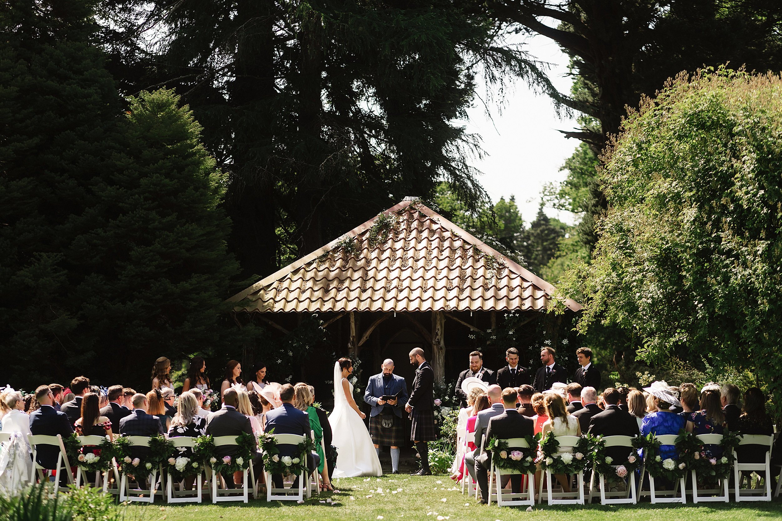 guests seated on white chairs in front of a wooden gazebo in the gardens of errol park wedding venue in scotland watch the officiant conduct the wedding ceremony for the bride and groom