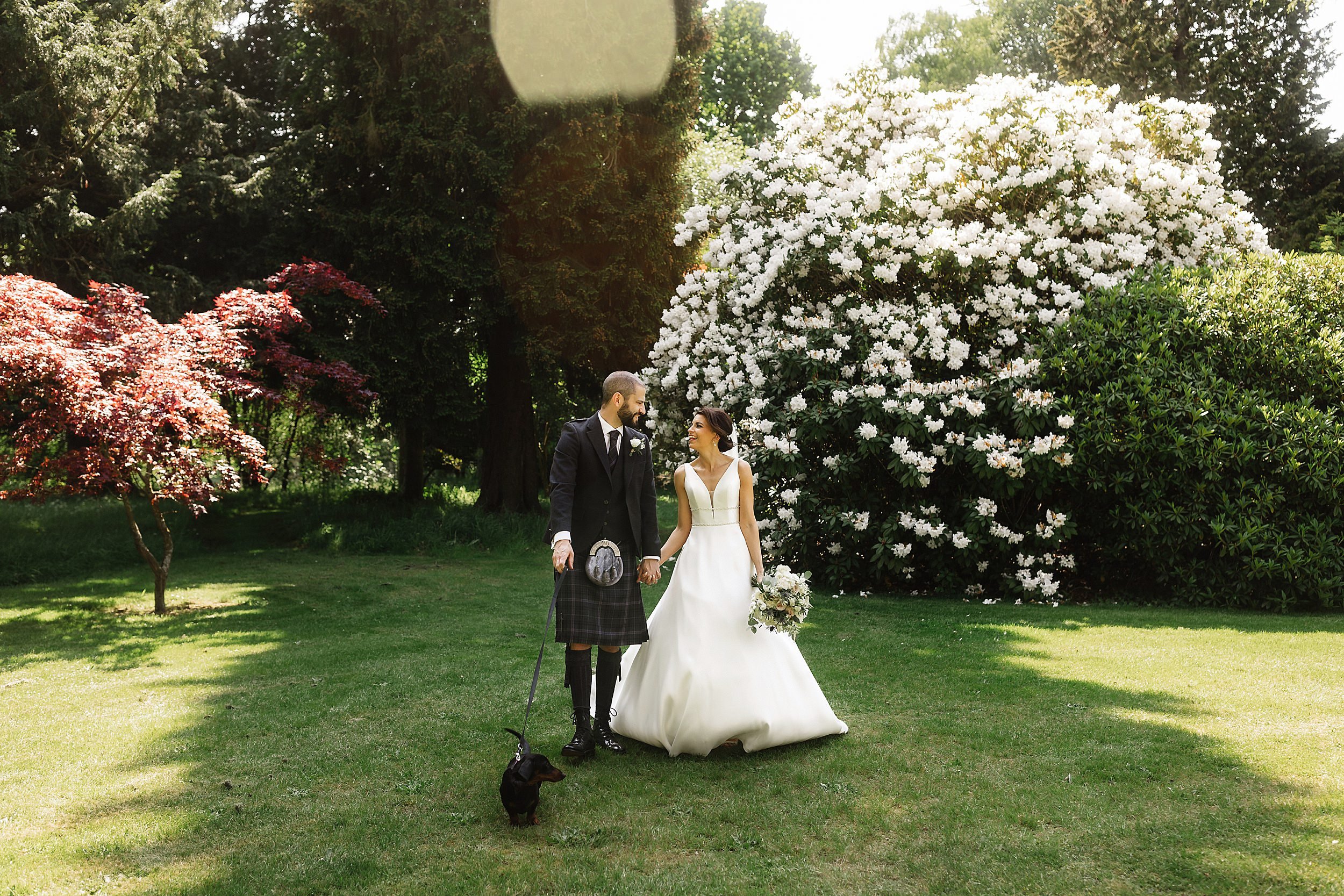 the bride and groom pose with their miniature daschund in the gardens of errol park wedding venue trees and flowering shrubs are visible in the background